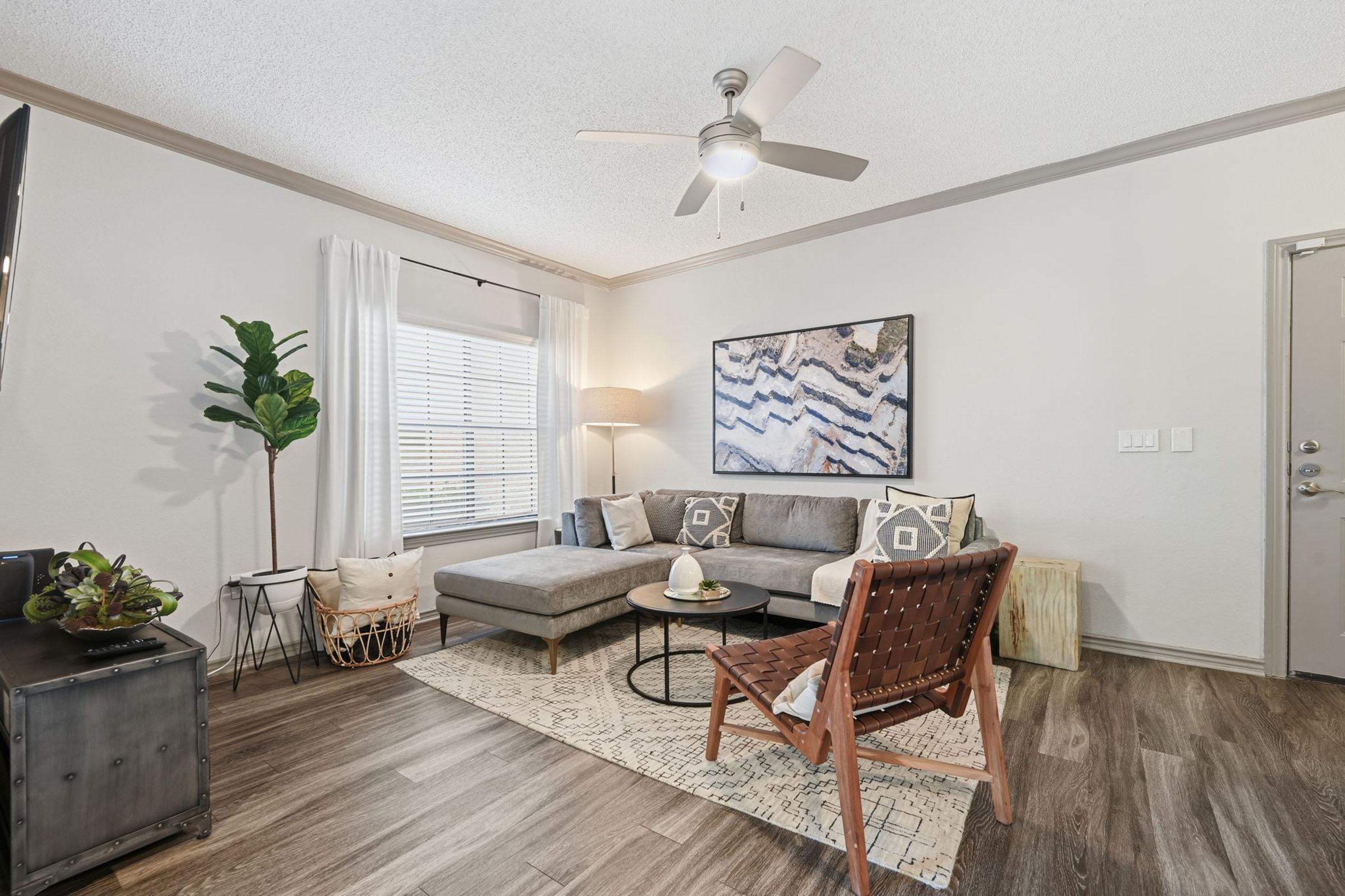 A cozy living room featuring a gray sectional sofa with decorative pillows, a rustic wooden coffee table, and a stylish chair. There's a ceiling fan overhead, a large window with sheer curtains, and a potted plant in the corner. The flooring is a warm wood laminate with a decorative rug adding texture.