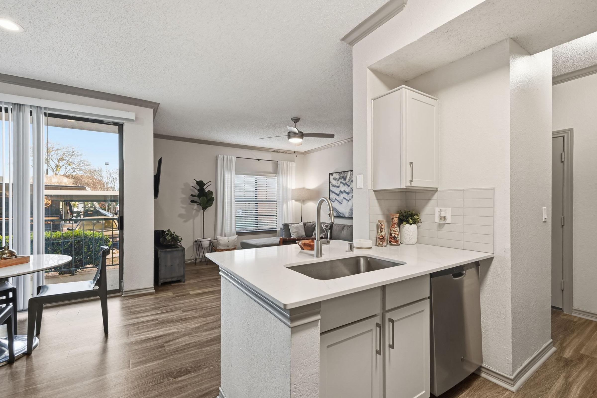 Modern kitchen featuring a white countertop, stainless steel sink, and light cabinetry. The open layout connects to a cozy living area with a fan and large windows, allowing natural light. A small dining table sits nearby, and there are decorative plants and items on display.