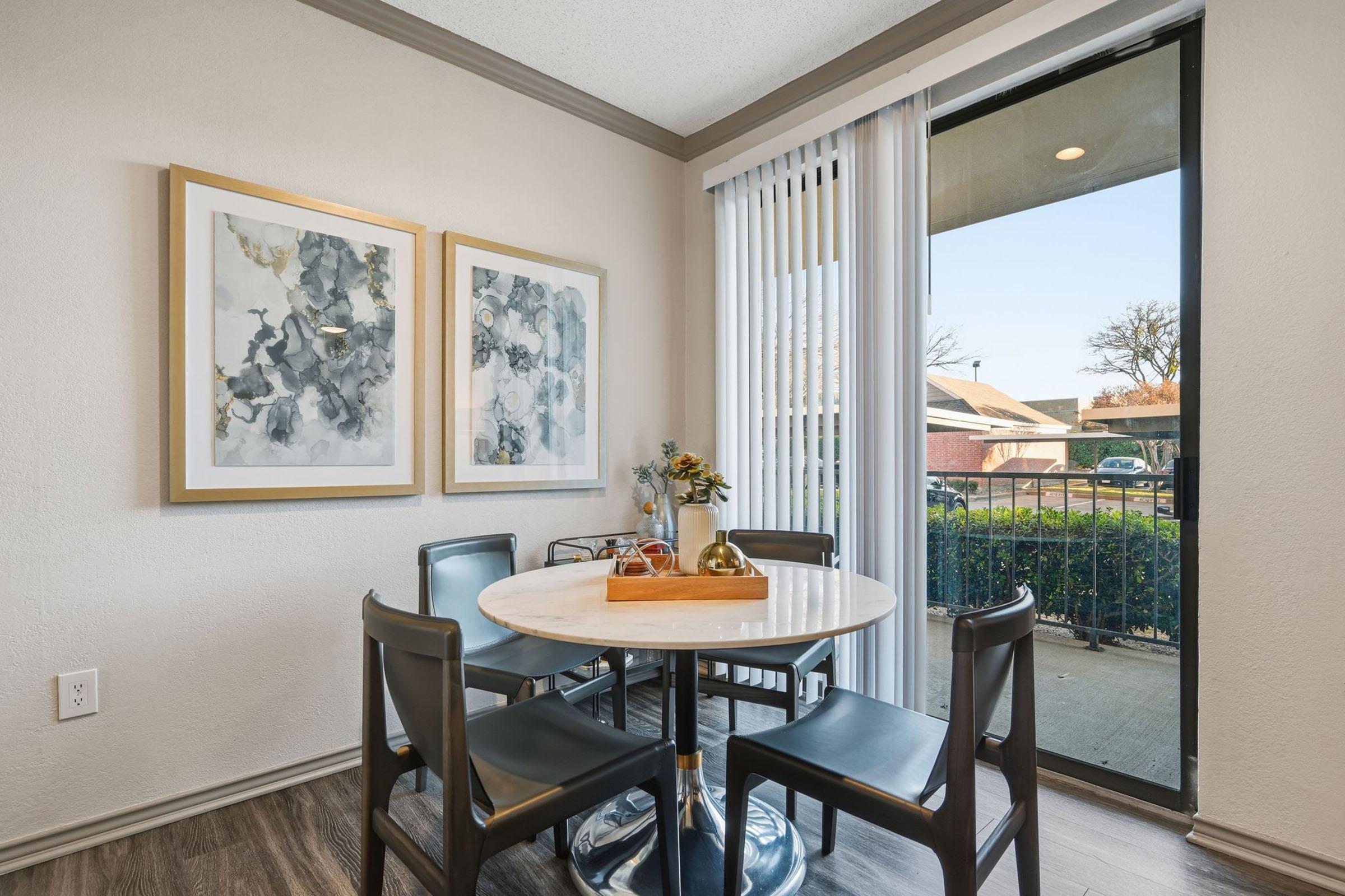 A cozy dining area featuring a round marble table surrounded by four black chairs. Two framed abstract art pieces adorn the wall, with large windows offering natural light and a view of the outdoors. The space has a warm and inviting atmosphere, ideal for meals or casual gatherings.