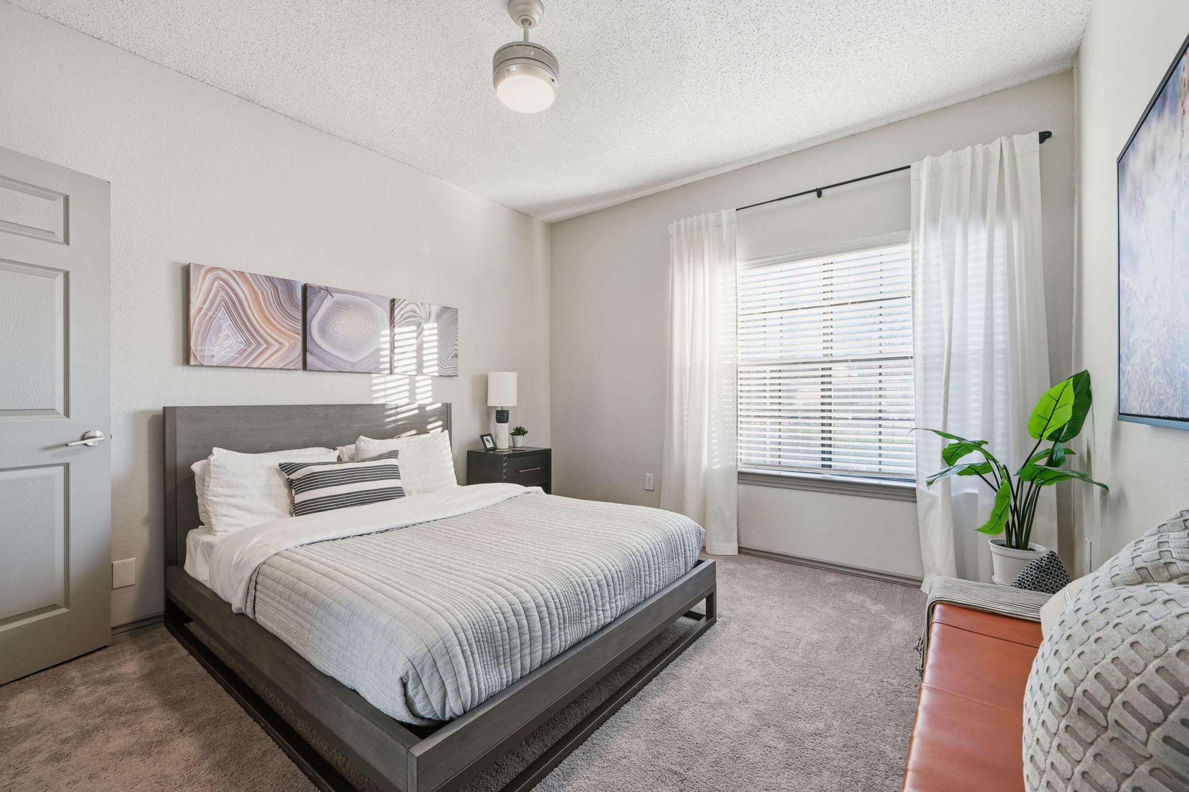 A cozy bedroom featuring a comfortable bed with light gray bedding, a decorative headboard, and a bedside table. The room has natural light streaming through a window with white curtains, a small plant in the corner, a striped decorative pillow, and soft, neutral-colored walls and carpet.