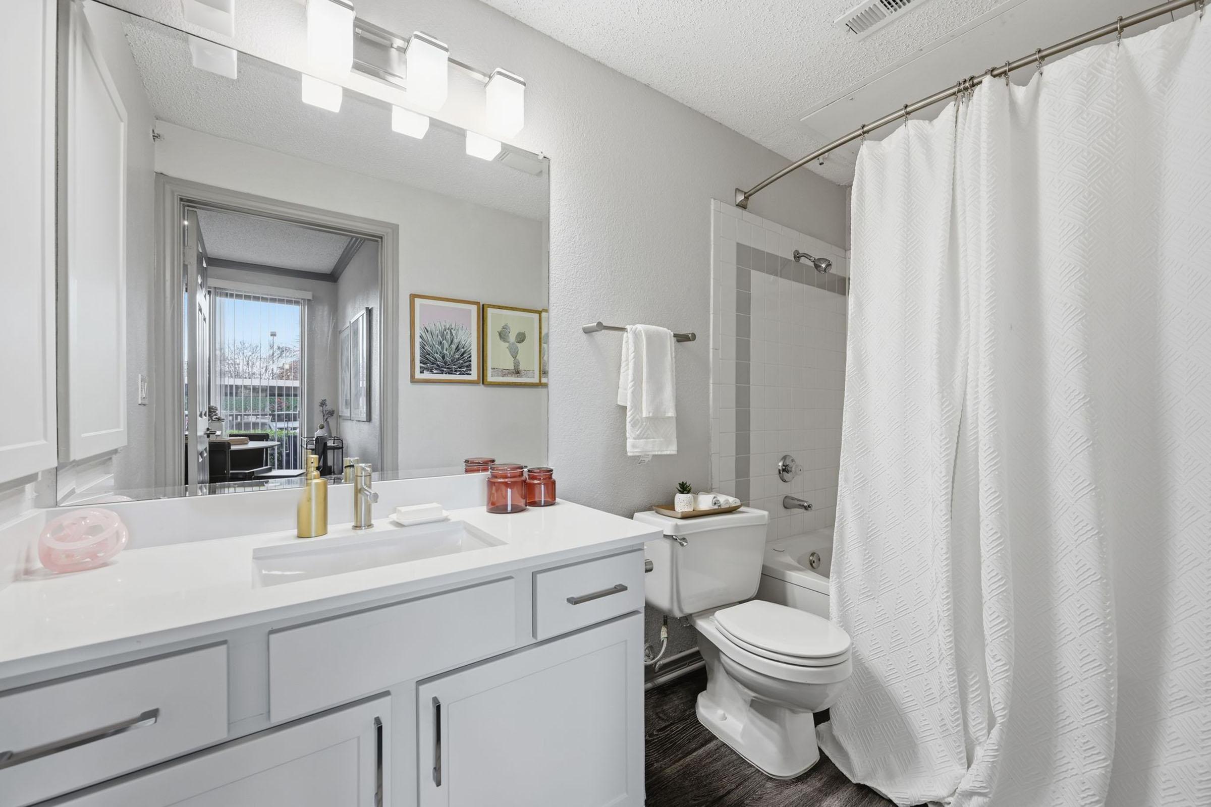 Modern bathroom with a white vanity featuring a gold faucet, large mirror, and two decorative frames on the wall. A white shower curtain separates the shower area, which includes a white tiled wall. The space has soft lighting and a window visible in the background, enhancing the bright ambiance.