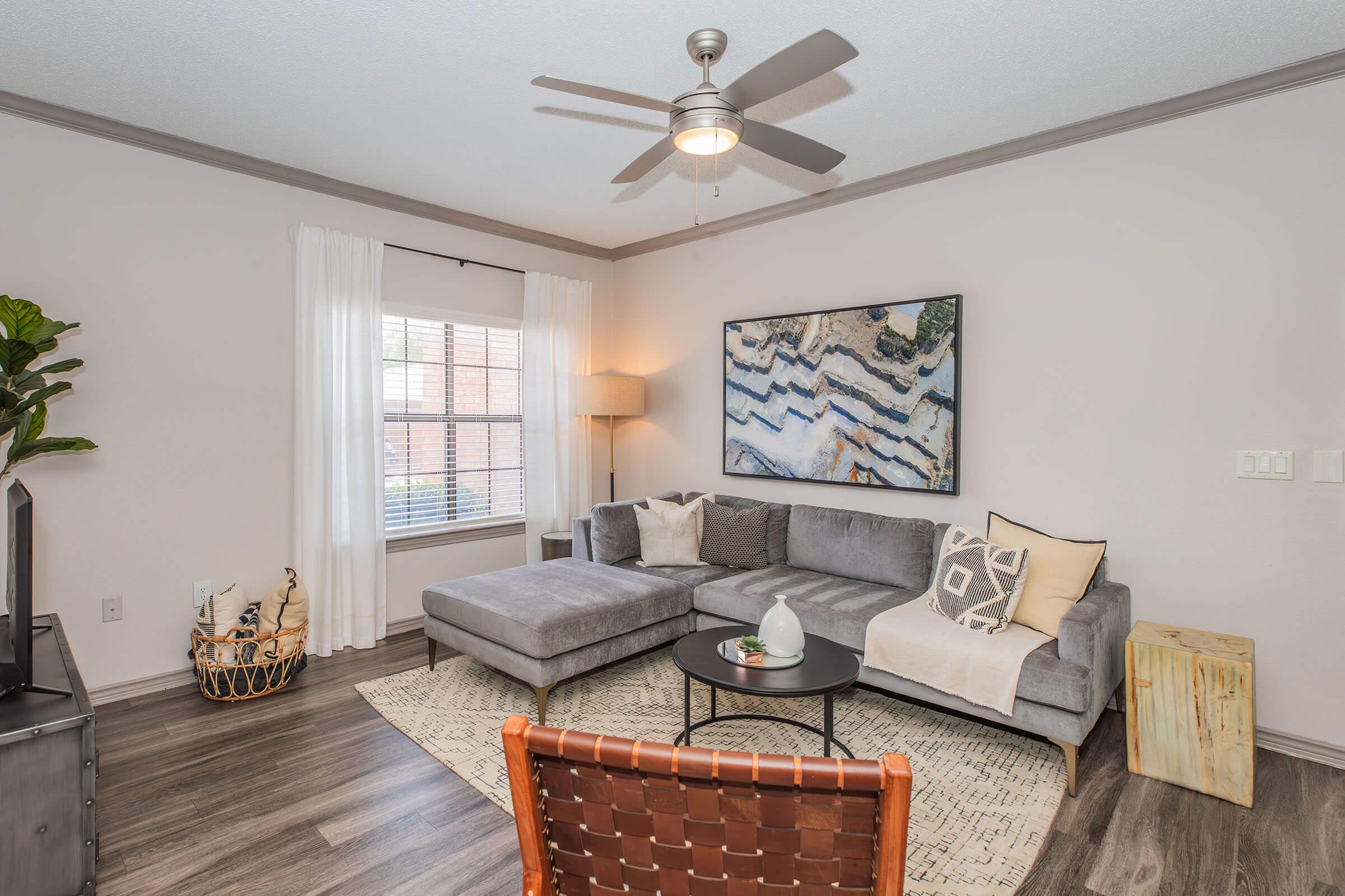 A cozy living room featuring a grey sectional sofa, a round coffee table, and a textured area rug. There's a large abstract painting on the wall, a lamp beside the sofa, and a decorative chair in the foreground. Natural light streams in through a window with white curtains, creating a warm and inviting atmosphere.