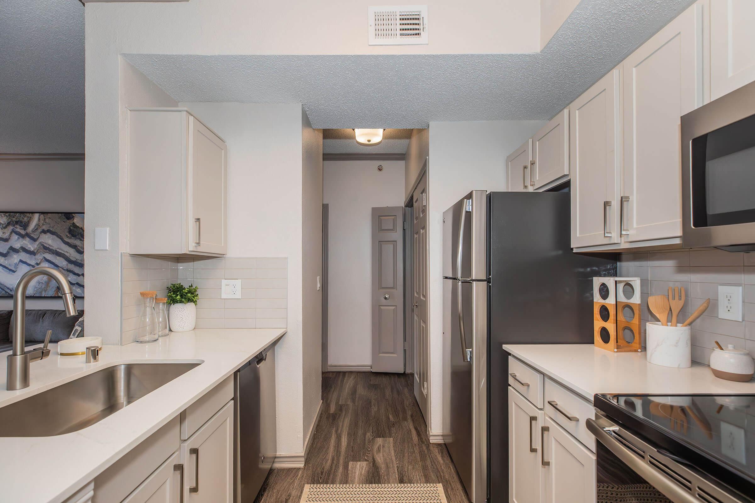 Modern kitchen featuring light-colored cabinetry, stainless steel appliances, and a window. The countertop is clean and organized with a few kitchen accessories, while the floor has a wood-like finish. A doorway leads to a hallway, enhancing the open layout. Natural light brightens the space.