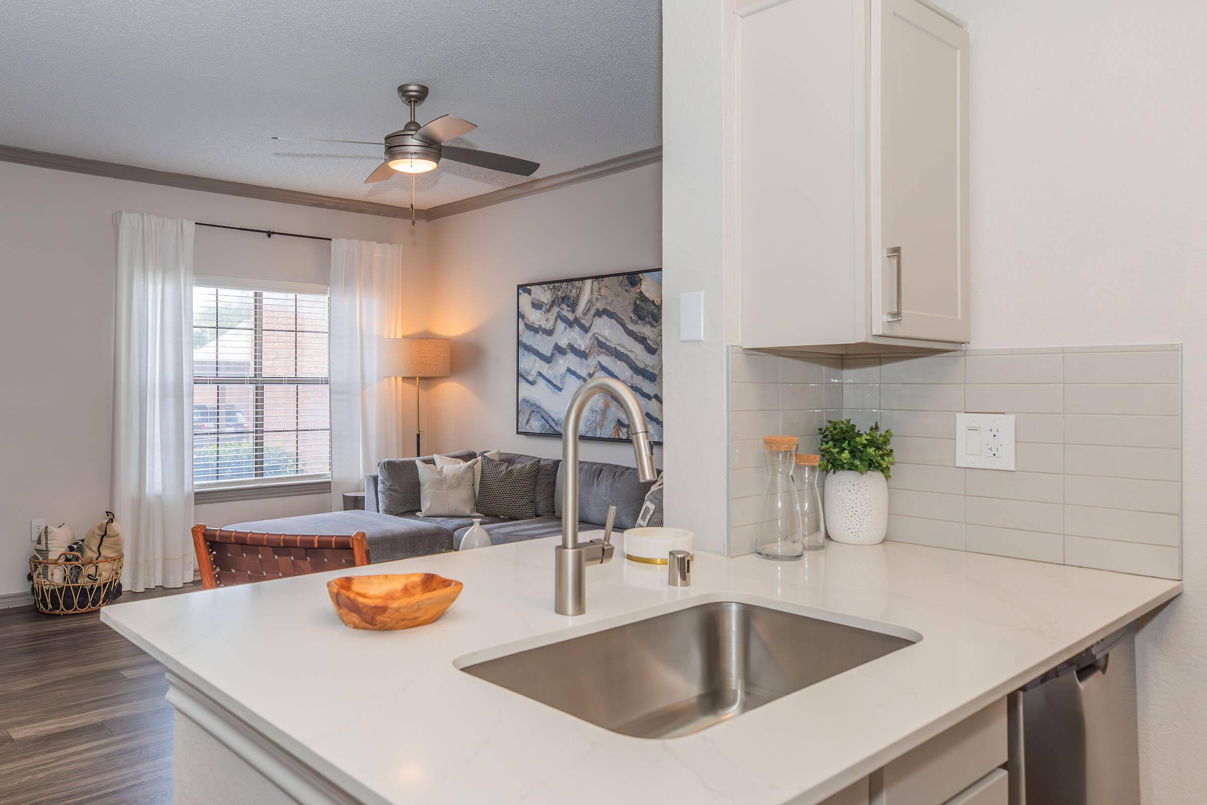 A modern kitchen with a stainless steel sink and light-colored countertop, featuring a bowl and kitchen accessories. In the background, a cozy living area with a gray sofa, a lamp, and a large window with white curtains. Decorative elements include a potted plant and wall art.