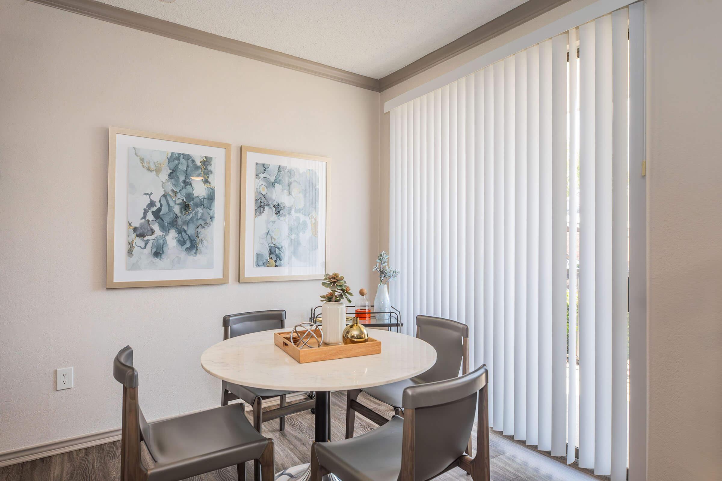 A cozy dining area featuring a round table surrounded by four gray chairs. Two framed abstract artworks adorn the light-colored wall. A wooden tray with decorative items sits on the table. Large vertical blinds allow natural light to filter in from a nearby window.