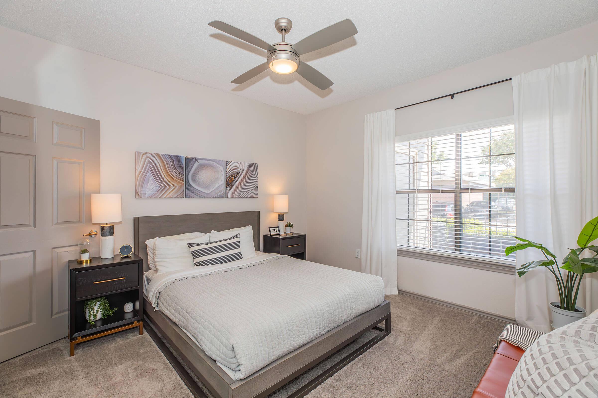 A cozy bedroom featuring a queen-sized bed with light bedding, two bedside tables with lamps, and a modern ceiling fan. The room has a large window with white curtains allowing natural light, showcasing a small plant and decorative artwork on the wall.