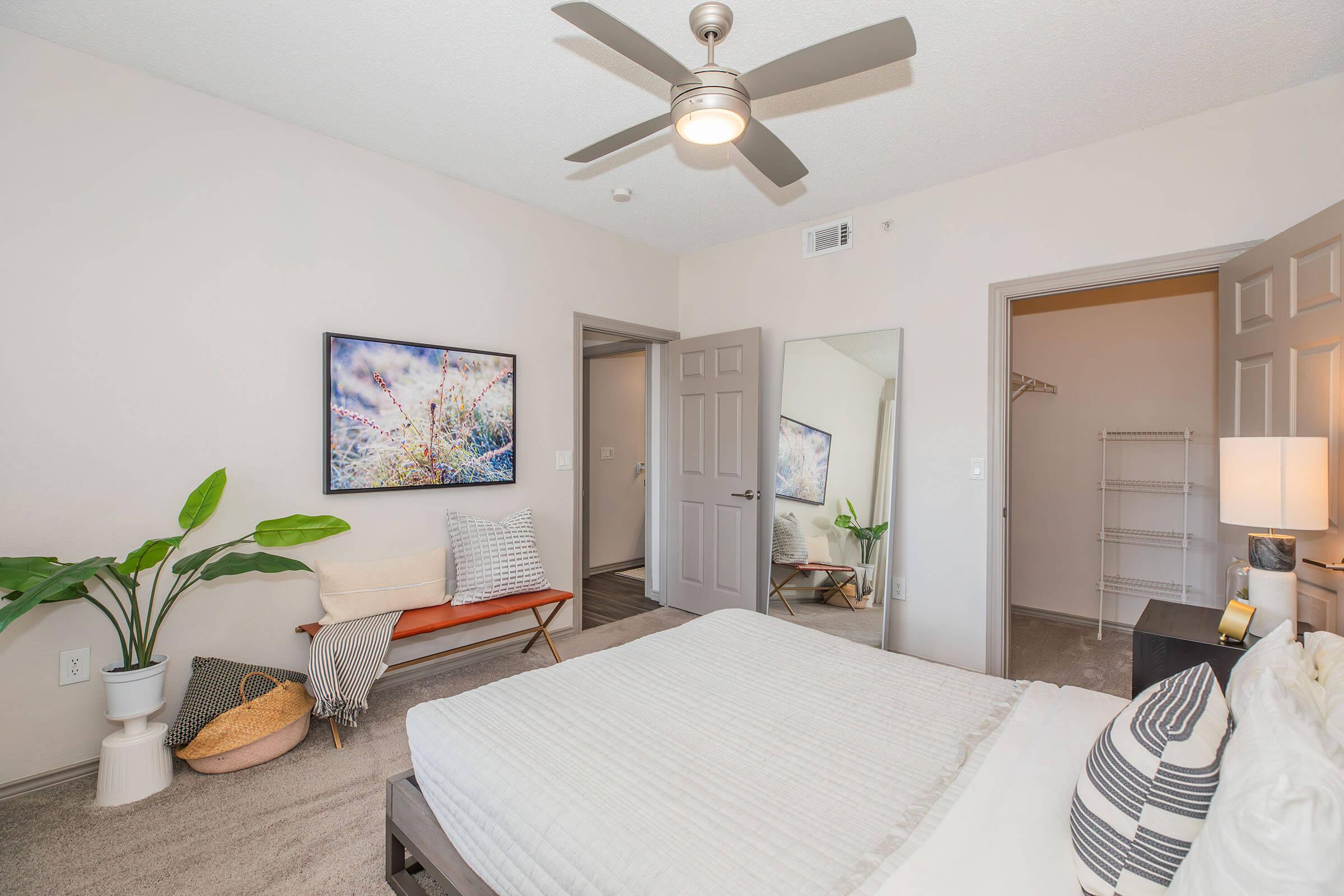 A cozy bedroom featuring a neatly made bed with white linens, a ceiling fan, and a large mirror. There are decorative elements like a plant, a piece of artwork on the wall, and a small bench with cushions. The room has light-colored walls and a warm, inviting atmosphere.