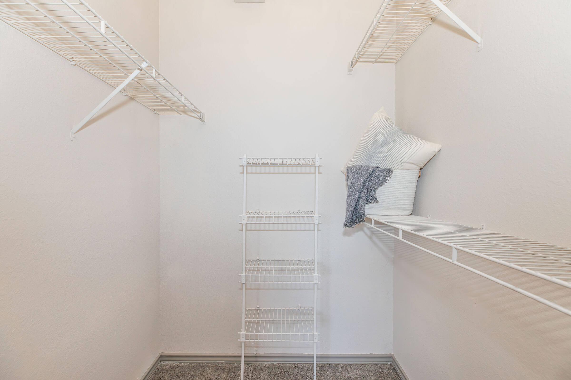 A spacious empty closet featuring white wire shelving on two walls. In the center, there's a metal shelving unit with no items on it. A decorative pillow rests on the floor, adding a cozy touch to the otherwise minimalist space with light-colored walls.