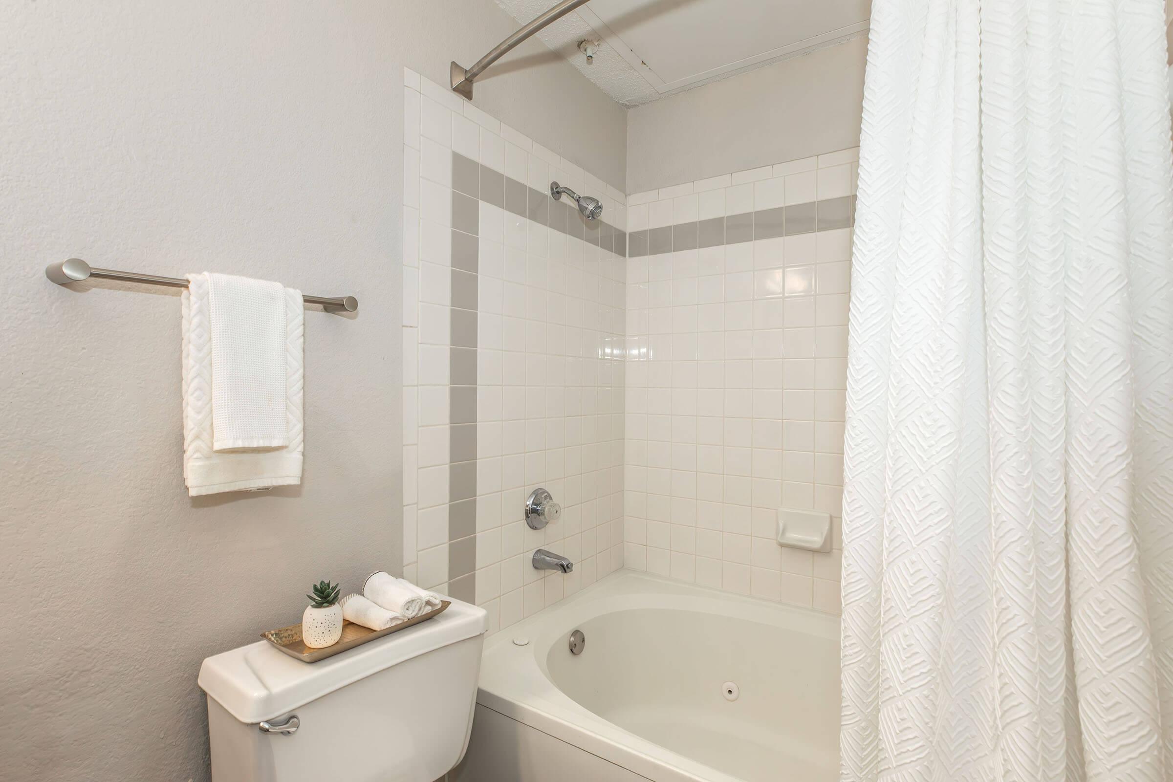A clean and modern bathroom featuring a white bathtub with a shower curtain, white tiled walls, a toilet with a decorative plant on top, and neatly arranged towels on a rack. The overall color scheme is neutral, creating a serene and inviting atmosphere.