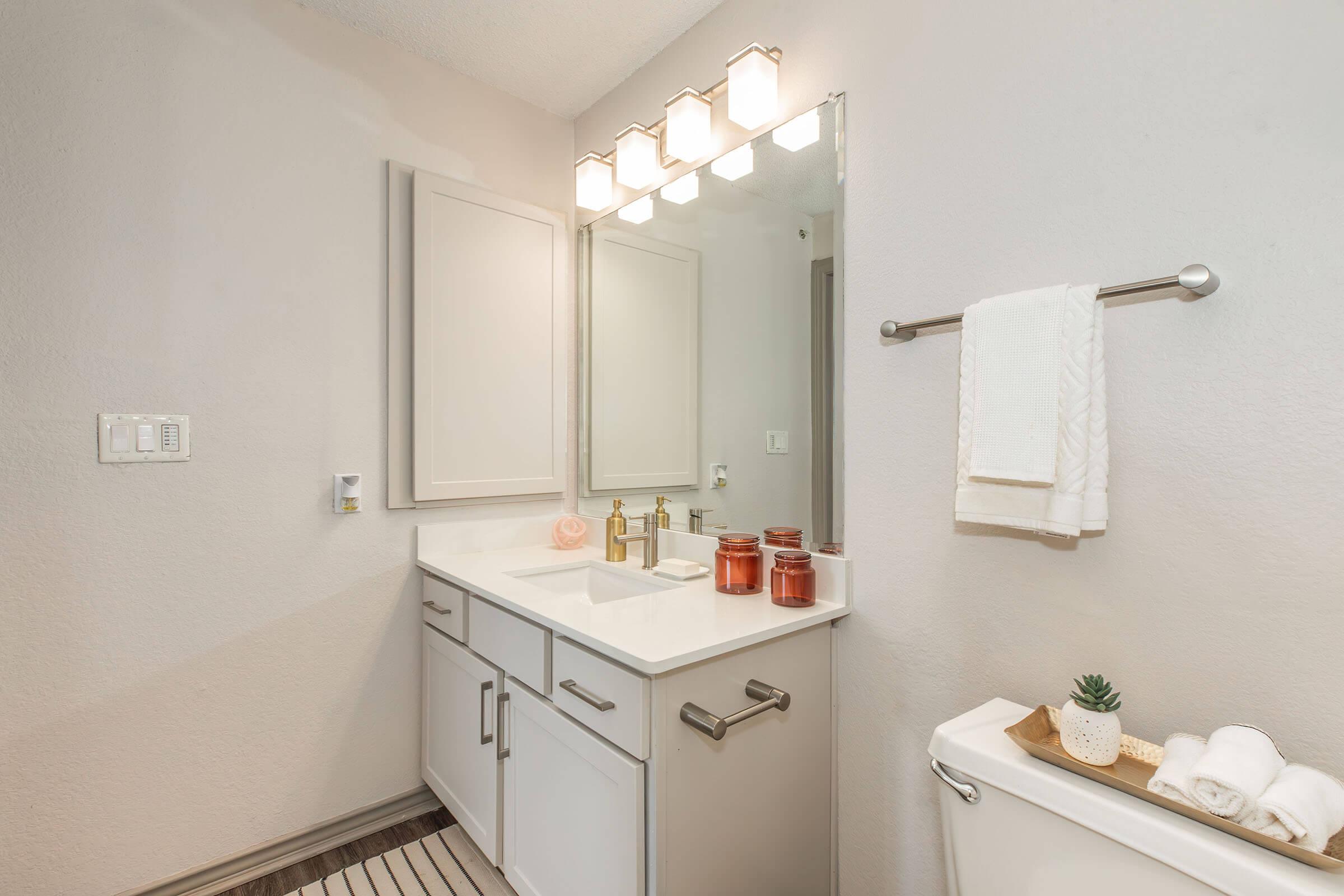 A modern bathroom featuring a white vanity with a sink, golden faucets, and a large mirror above it. The countertop holds decorative candles and a small plant. A towel rack with a rolled towel is attached to the wall, and a bathtub is visible in the background. Soft lighting illuminates the space.