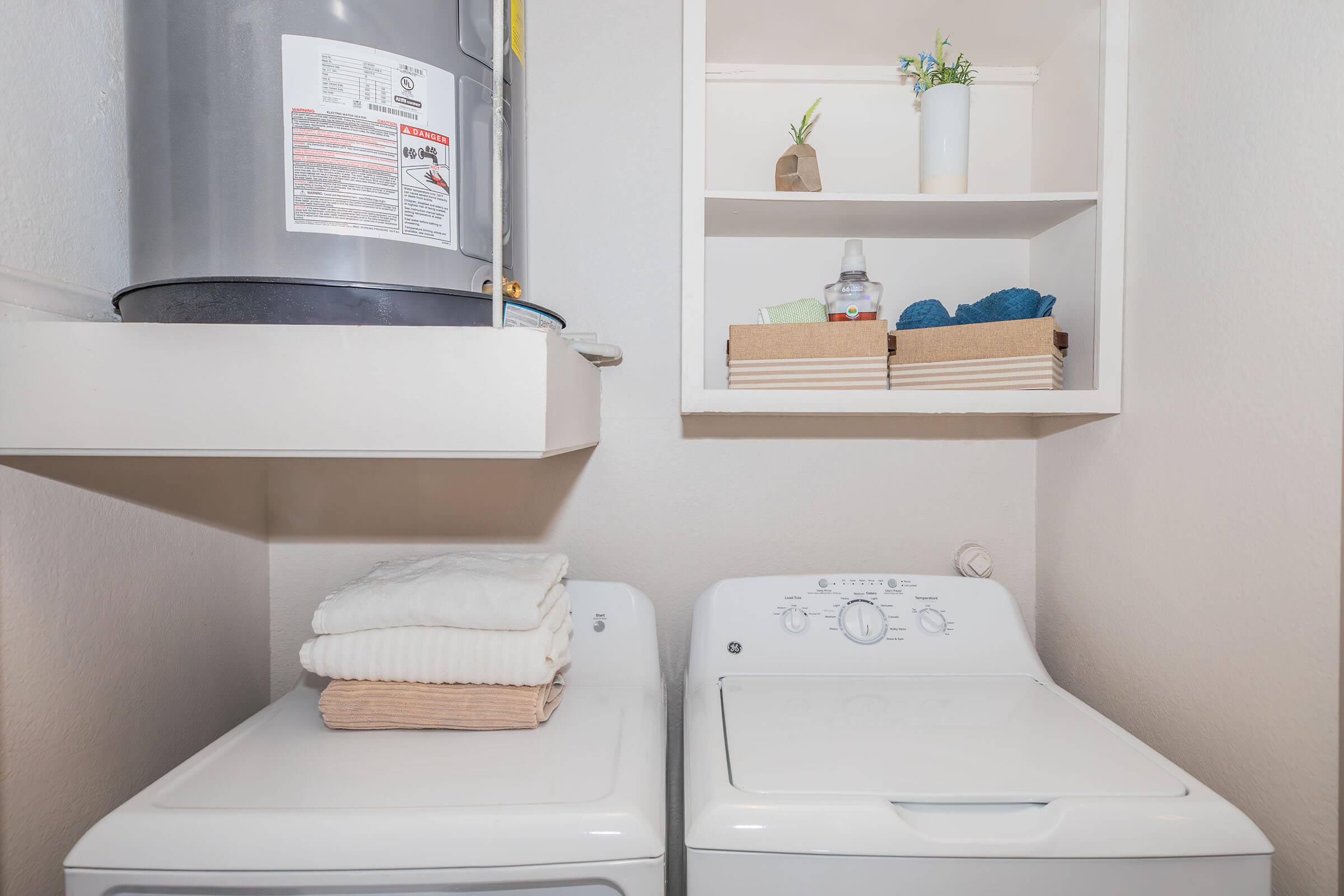 A laundry area featuring a white washing machine stacked with neatly folded towels, a dryer, and a shelf above displaying small potted plants and baskets containing various items. A water heater is visible on the wall.