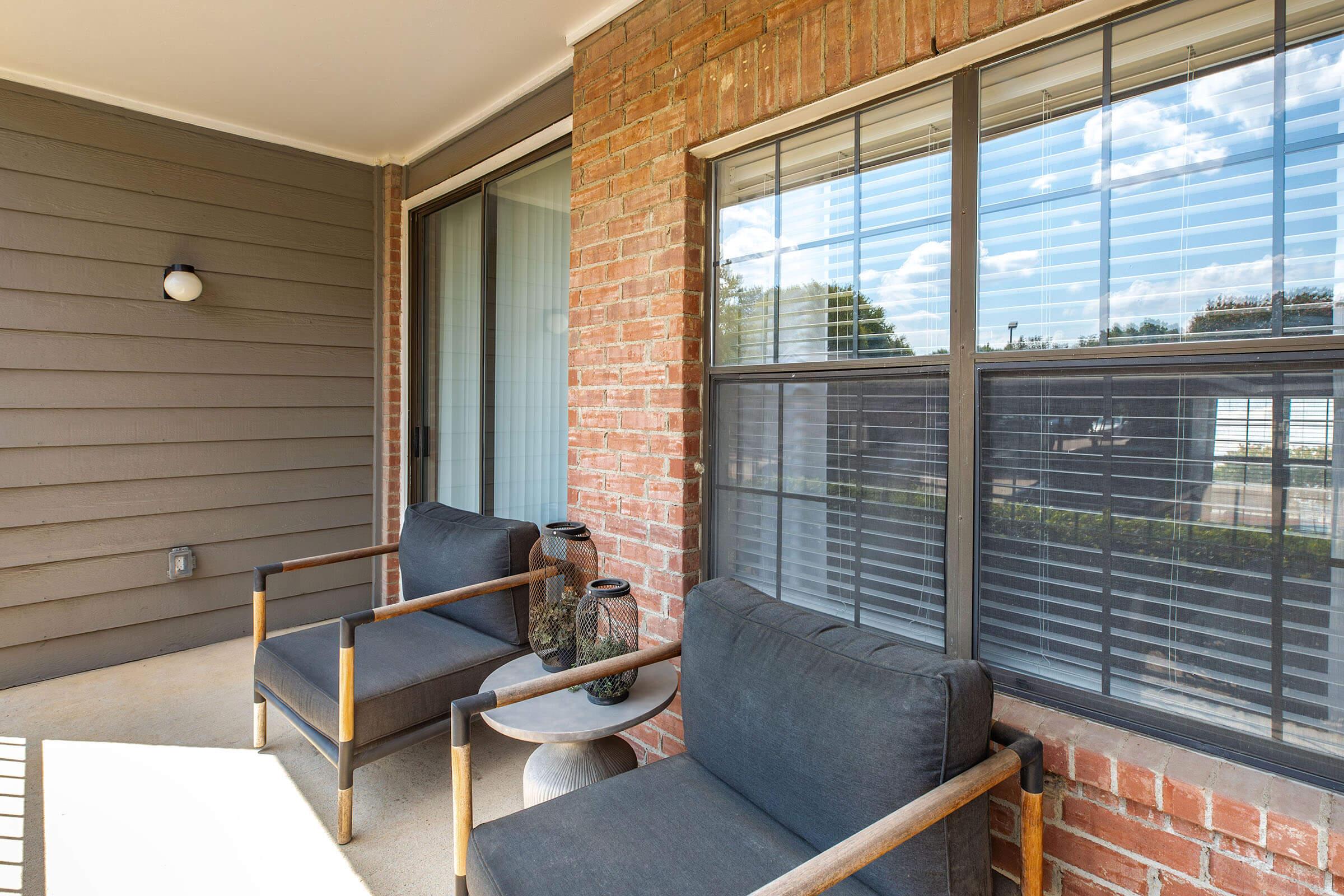 A cozy outdoor space featuring two black armchairs on a concrete patio, complemented by a decorative vase. The backdrop includes a brick wall with large windows, allowing plenty of natural light, and a light fixture on the side. Soft clouds are visible in the blue sky outside.