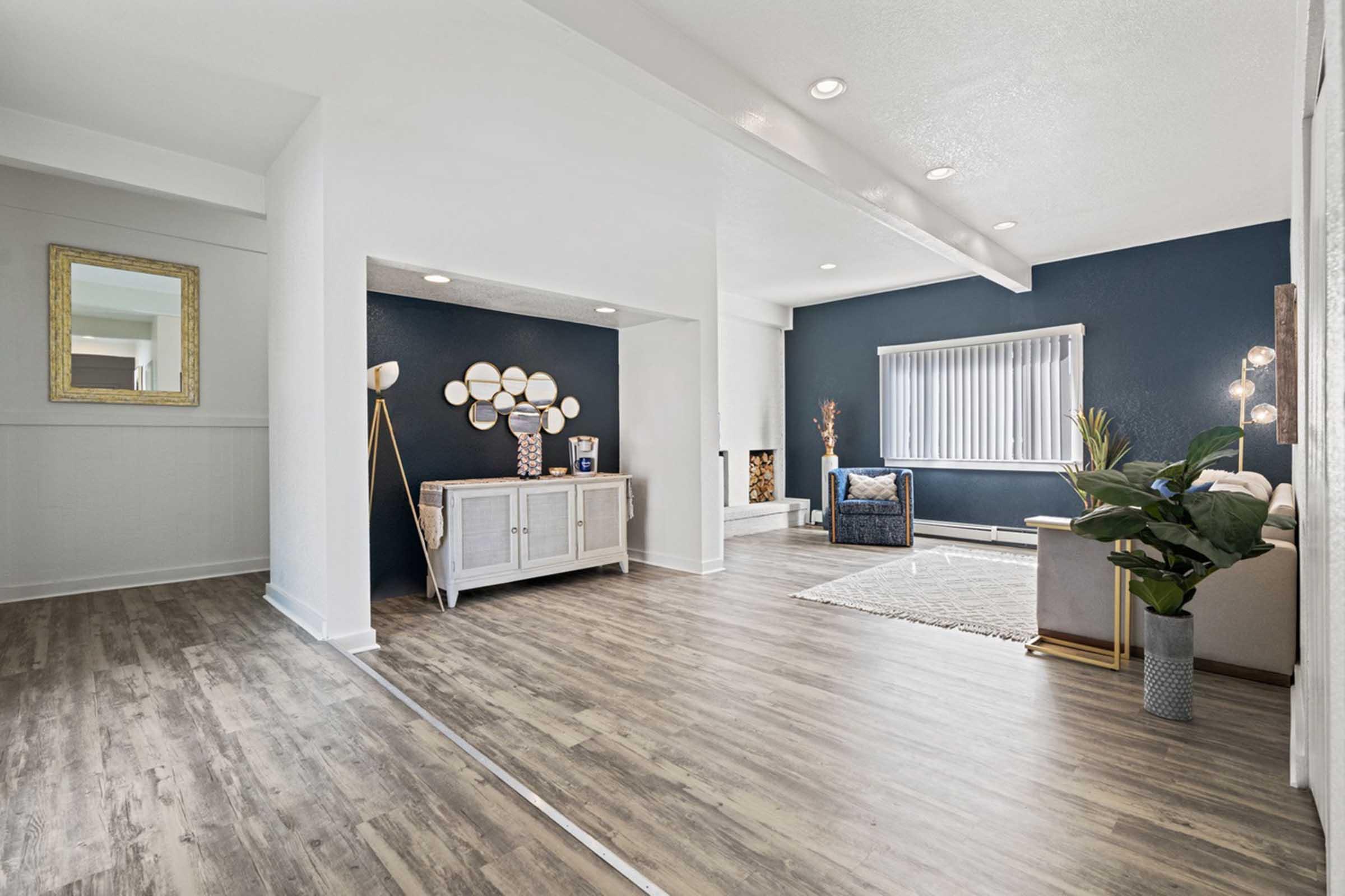 Bright and airy living space featuring modern decor. A stylish white console table stands against a dark blue accent wall, adorned with decorative elements. Natural light streams through large windows, illuminating a cozy seating area with a plant and a rug on the wooden floor, creating a welcoming atmosphere.