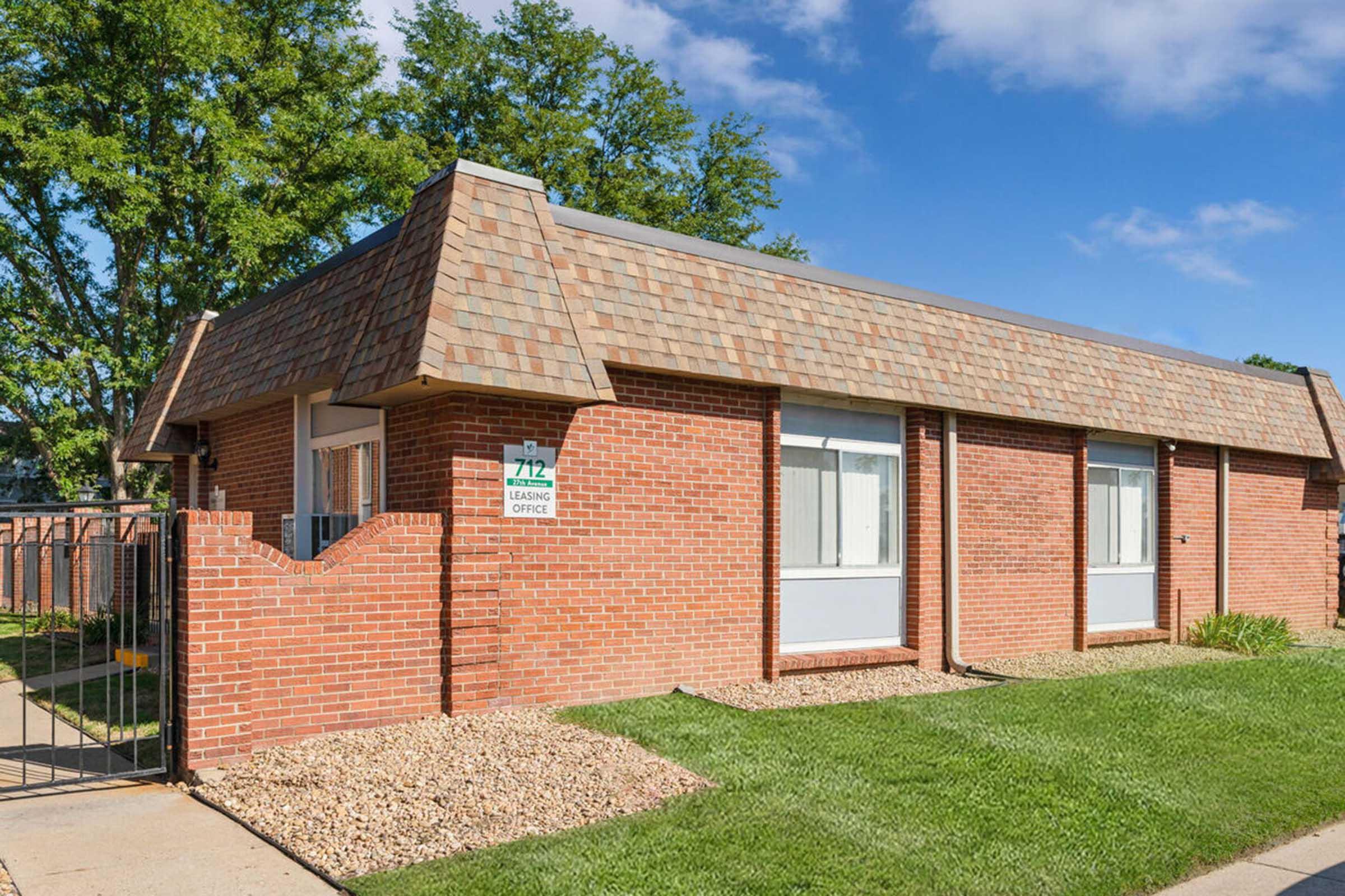 Single-story brick building with a sloped, shingled roof. The building features a sign indicating it is a leasing office. Well-maintained green lawn and small gravel area in front. Clear blue sky and trees in the background provide a pleasant atmosphere.