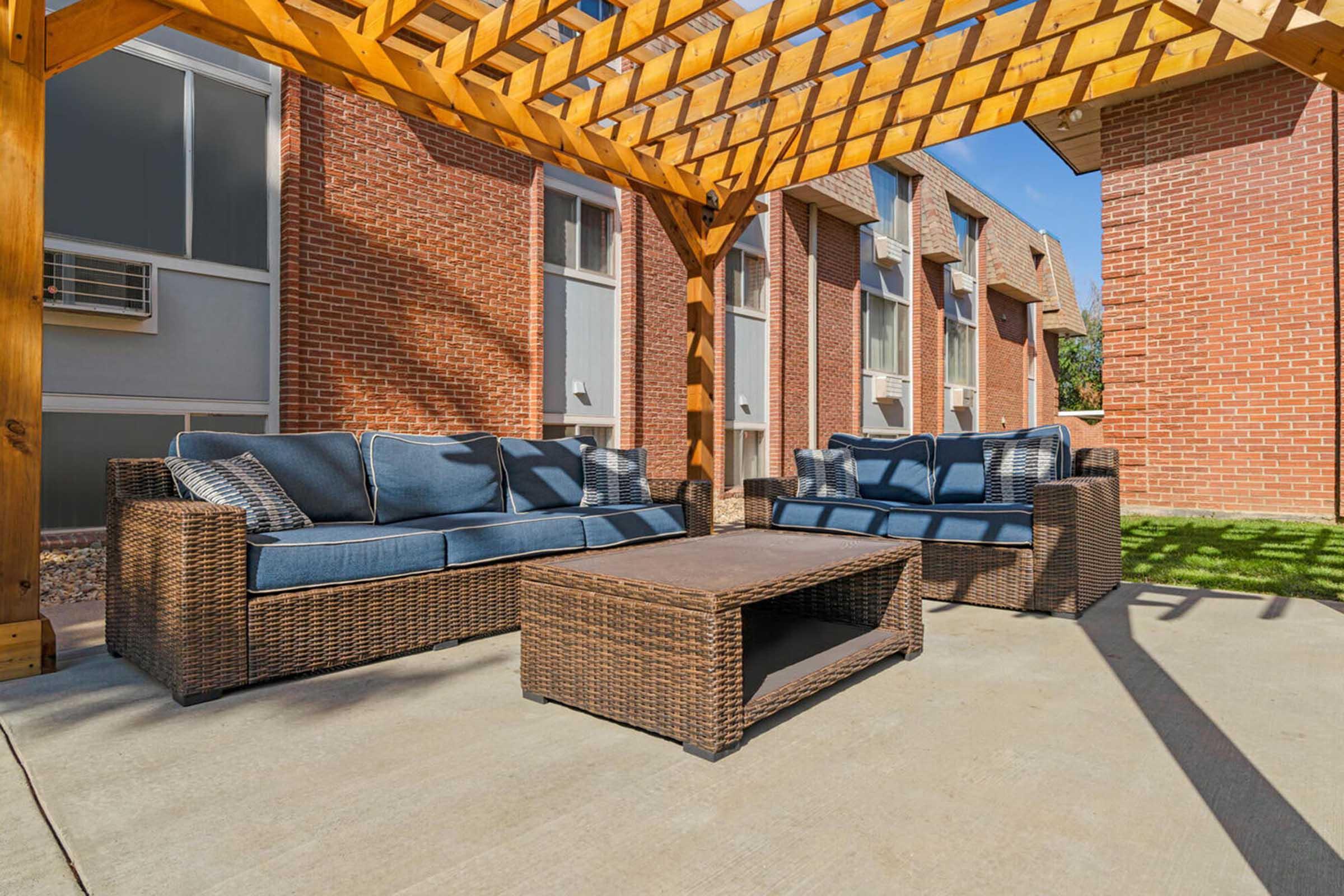 Outdoor seating area featuring two blue cushioned sofas and a coffee table under a wooden pergola. The background shows brick buildings and a patch of grass, creating a cozy, inviting atmosphere for relaxation or social gatherings.