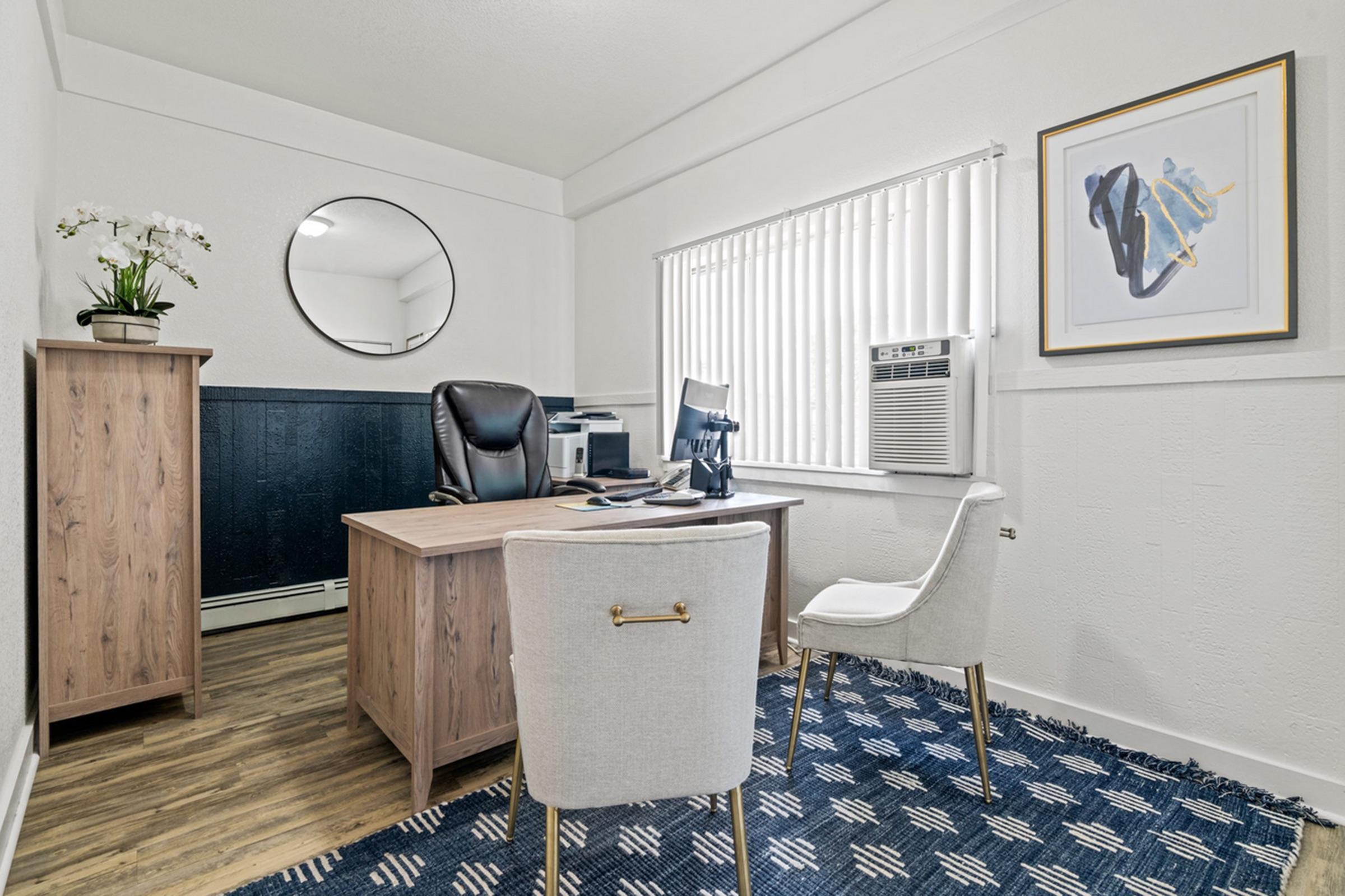 A modern office space featuring a wooden desk with a black office chair, a white chair, and an air conditioning unit. There is a round mirror on the wall, a tall plant on the desk, and framed artwork nearby. The floor is covered with a blue-patterned rug, and natural light enters through the window with vertical blinds.