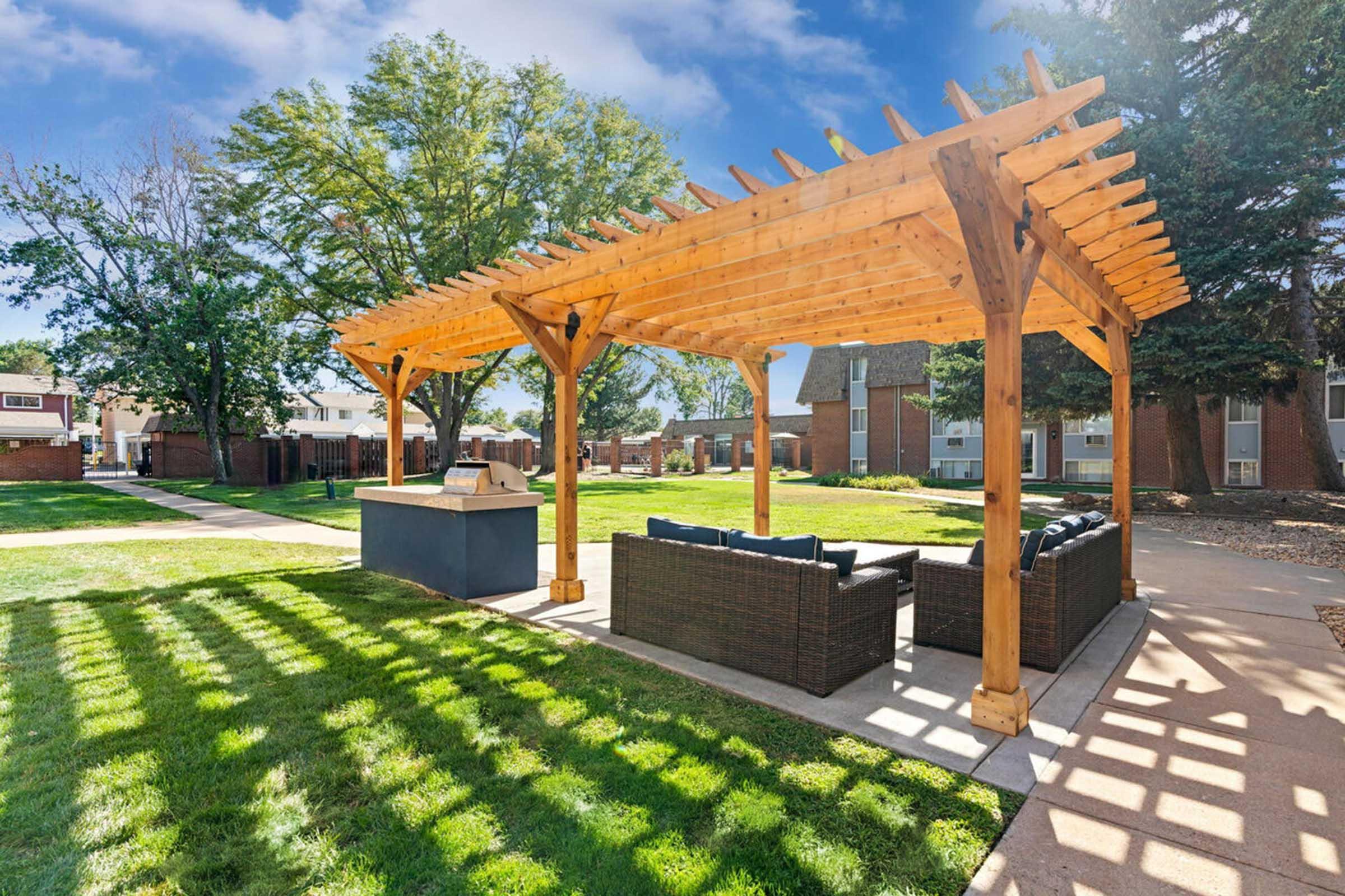 A well-maintained outdoor space featuring a wooden pergola with seating, surrounded by green grass and trees. The area includes a barbecue grill under the pergola and paved pathways, creating a welcoming environment for relaxation and social gatherings.