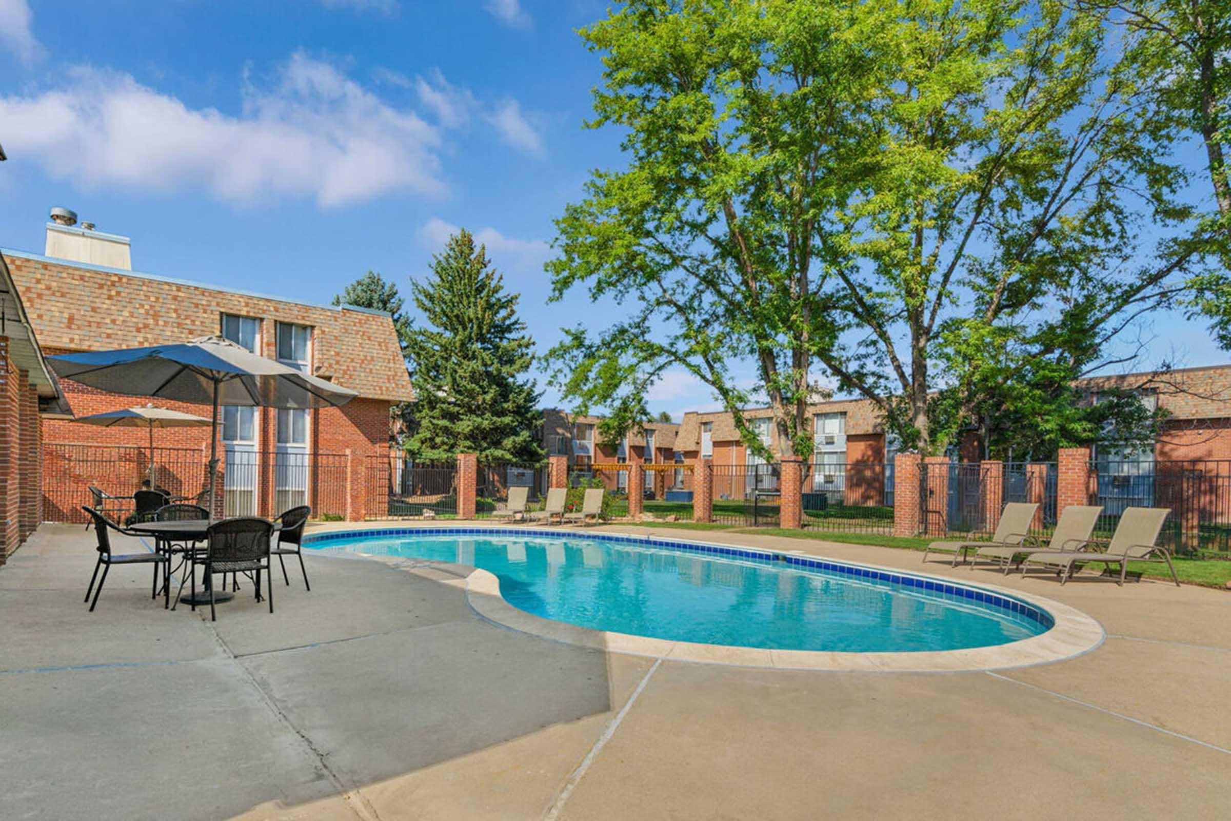 A swimming pool surrounded by lounge chairs and an umbrella, set in a well-landscaped area with green trees and buildings in the background. The sky is clear with a few clouds, creating a bright and inviting atmosphere.