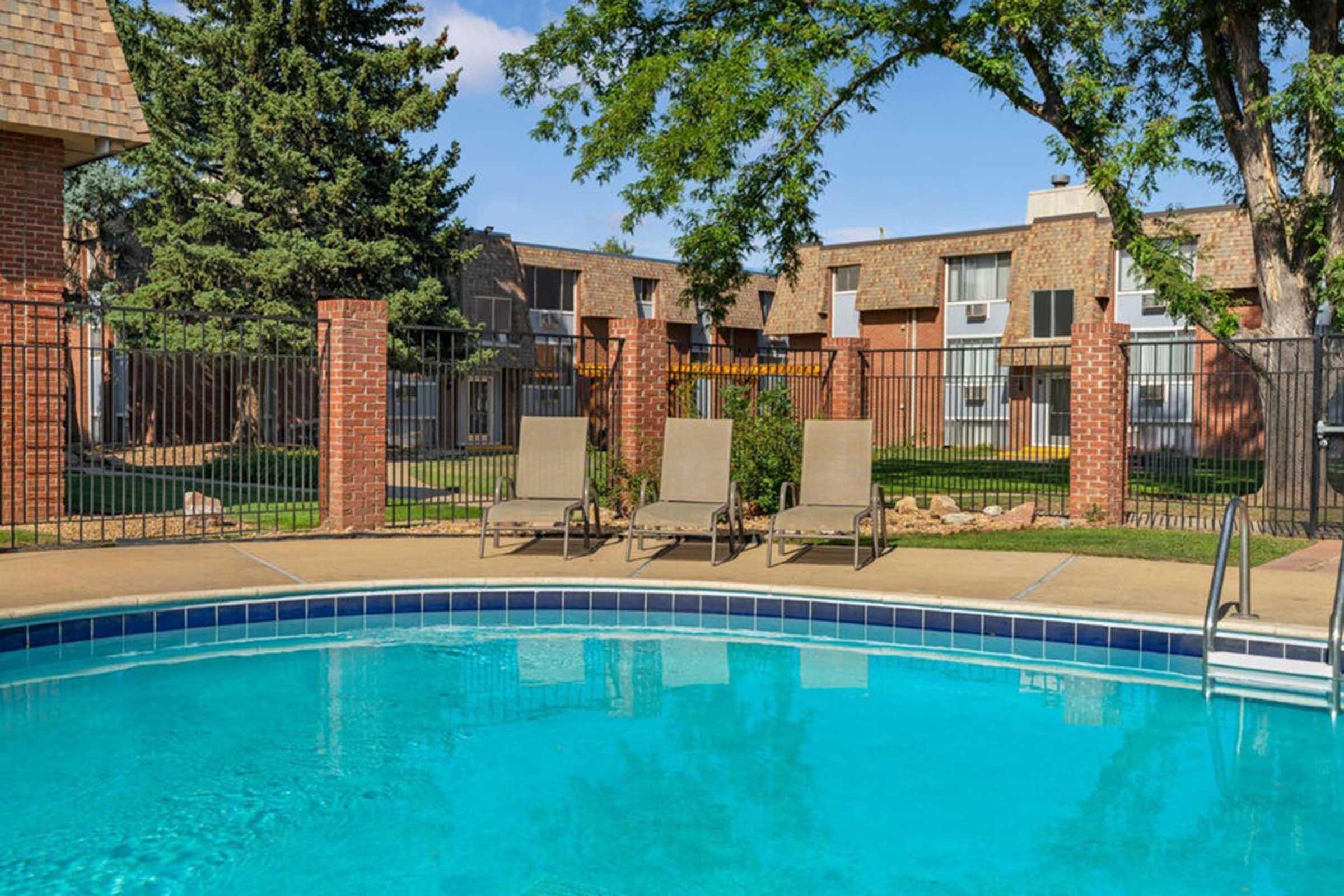 A clear swimming pool with blue water in the foreground, surrounded by a tan concrete deck. Three lounge chairs are positioned near the pool. In the background, there are brick buildings with balconies, greenery, and a fence enclosing the area, suggesting a residential community with outdoor amenities.