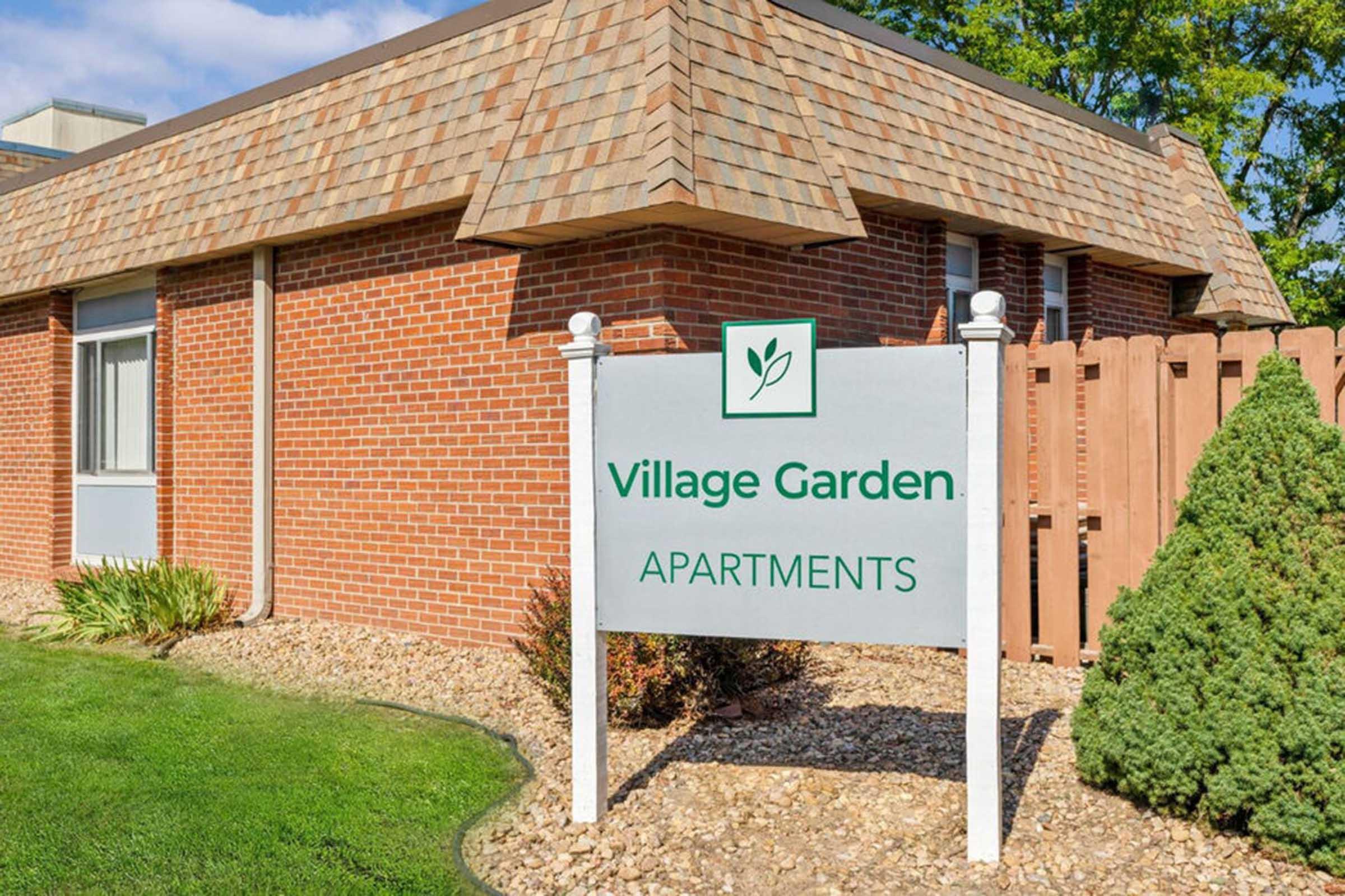 Sign for Village Garden Apartments in front of a brick building with a sloped roof and a landscaped area. The sign features a leaf logo and is surrounded by greenery and a wooden fence. The building is set against a blue sky with a few clouds.