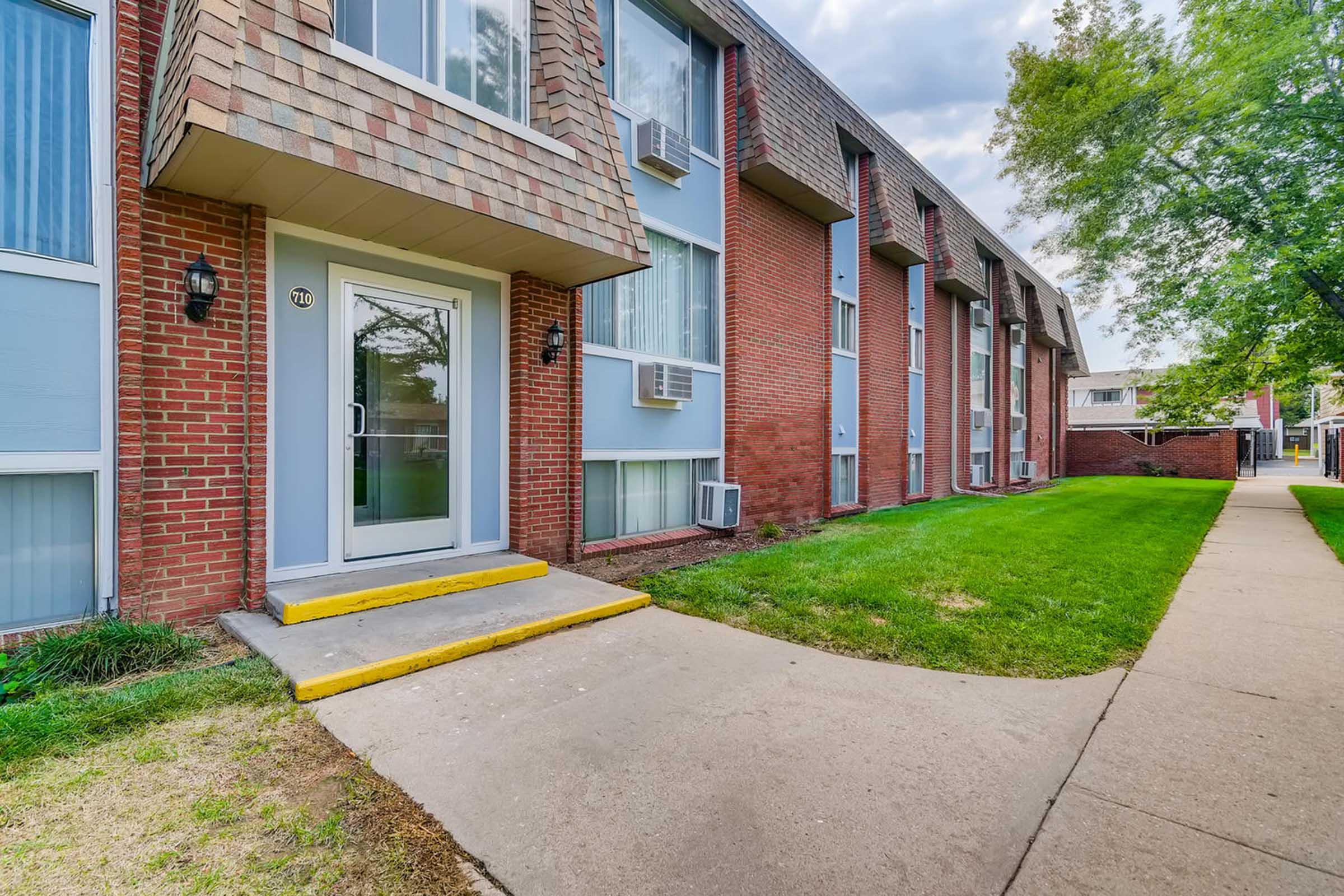 Exterior view of a multi-unit apartment building with a brick facade and large windows. A concrete pathway leads to the main entrance, which has a glass door and yellow steps. Air conditioning units are visible in some windows, and grassy areas are present on either side of the pathway. Trees line the background.