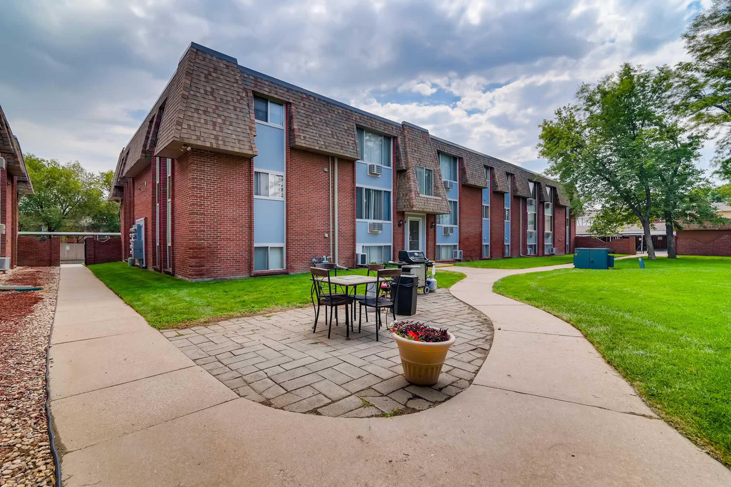 A view of a brick apartment complex featuring two buildings with large windows. In the foreground, there's a stone pathway leading to a small outdoor seating area with a table and chairs, surrounded by green grass and trees. The sky is partly cloudy, adding a pleasant ambiance to the scene.