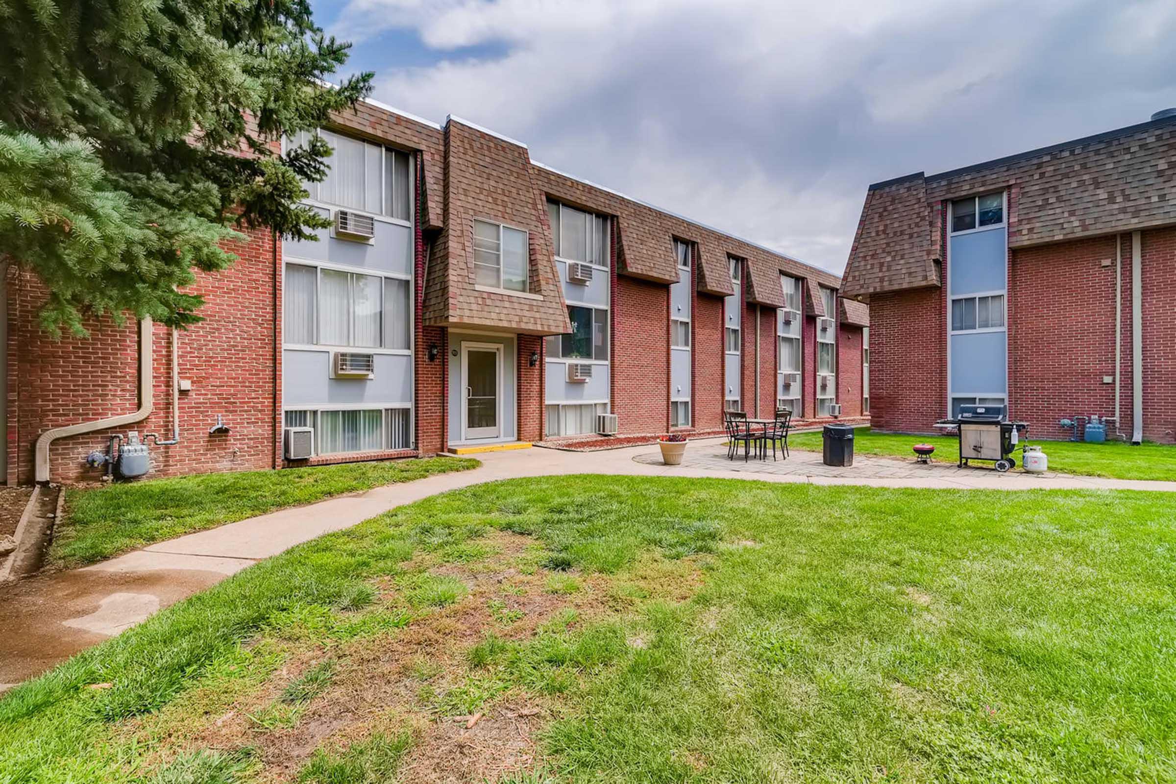 A view of a brick apartment building with multiple windows and air conditioning units. The landscaped area features a green lawn, a walking path, and outdoor furniture. In the background, another building is visible, highlighting a typical residential complex setup. The sky is partly cloudy.