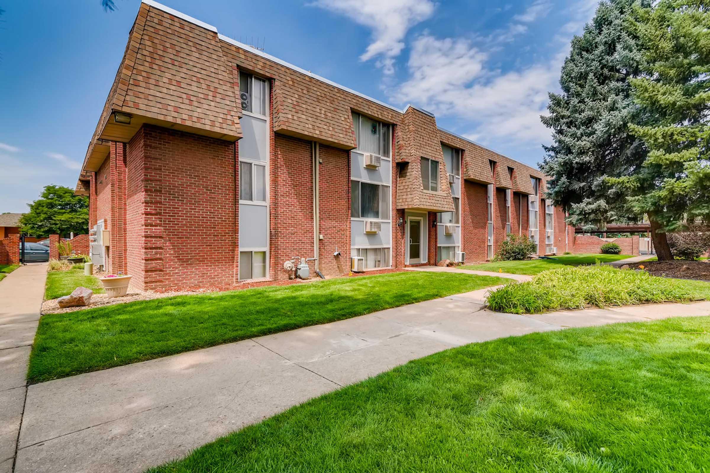 A red brick apartment building with multiple windows and air conditioning units. The building features a well-maintained lawn with green grass and shrubs. A concrete pathway leads to the entrance, and tall trees are visible in the background under a partly cloudy sky.