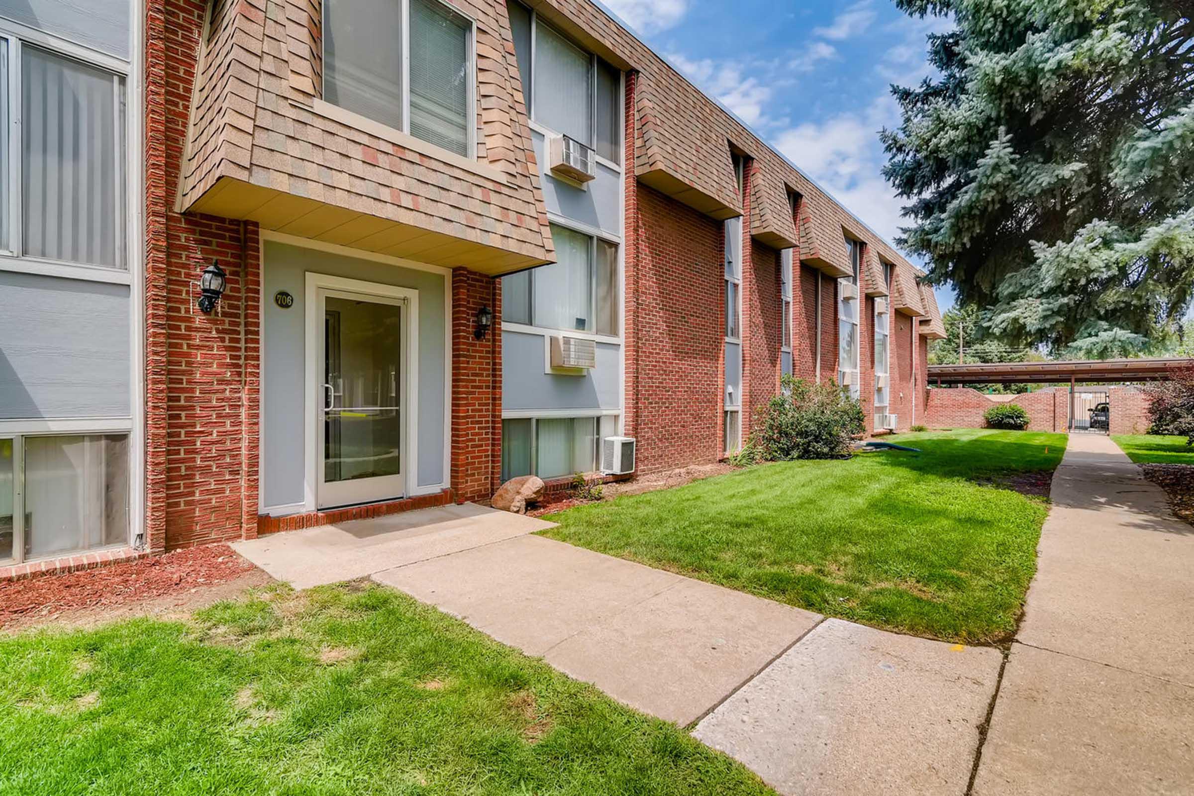 Exterior view of a brick apartment building with multiple windows and air conditioning units. A concrete walkway leads to the entrance, surrounded by well-maintained grass. There are shrubs and trees in the vicinity, under a bright blue sky with a few clouds.