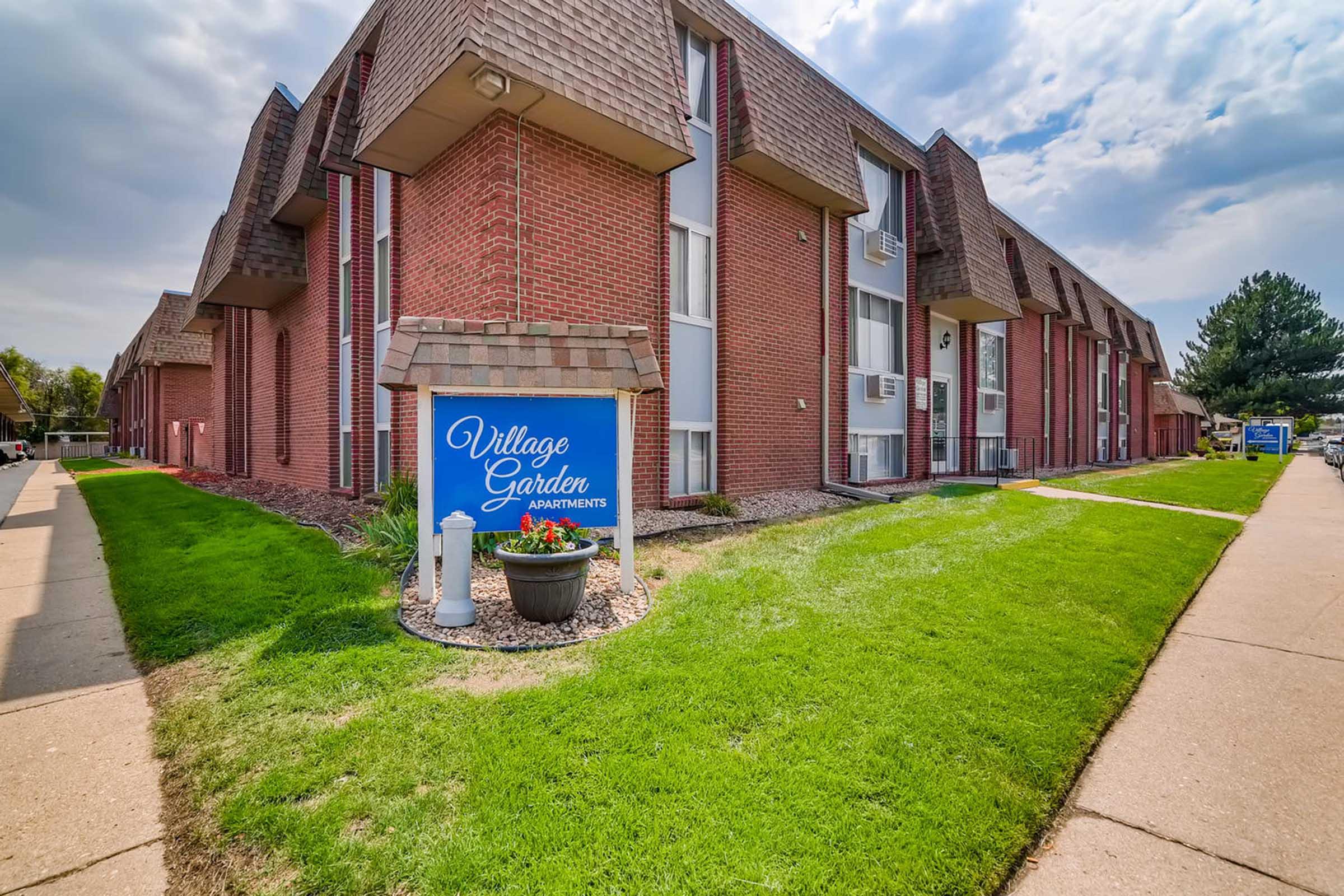 Exterior view of Village Garden Apartments, featuring a brick building with multiple floors, a sign in front, and well-maintained green grass and landscaping. The sky is partly cloudy, and the area is designed for residential living with walkways leading to the entrance.