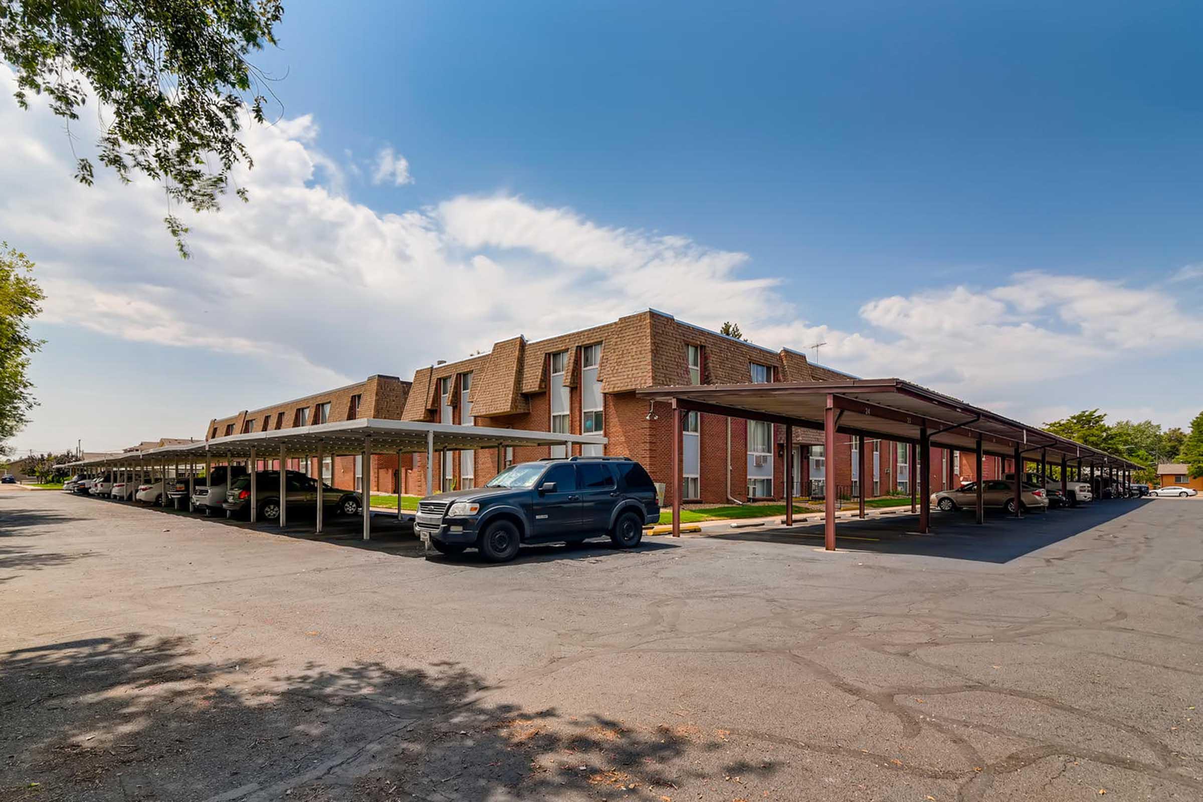A view of a row of buildings with a mix of one- and two-story structures under a clear blue sky. Covered parking spaces line the front, with several parked cars. The area is well-maintained, featuring green grass and trees, creating a pleasant residential environment.