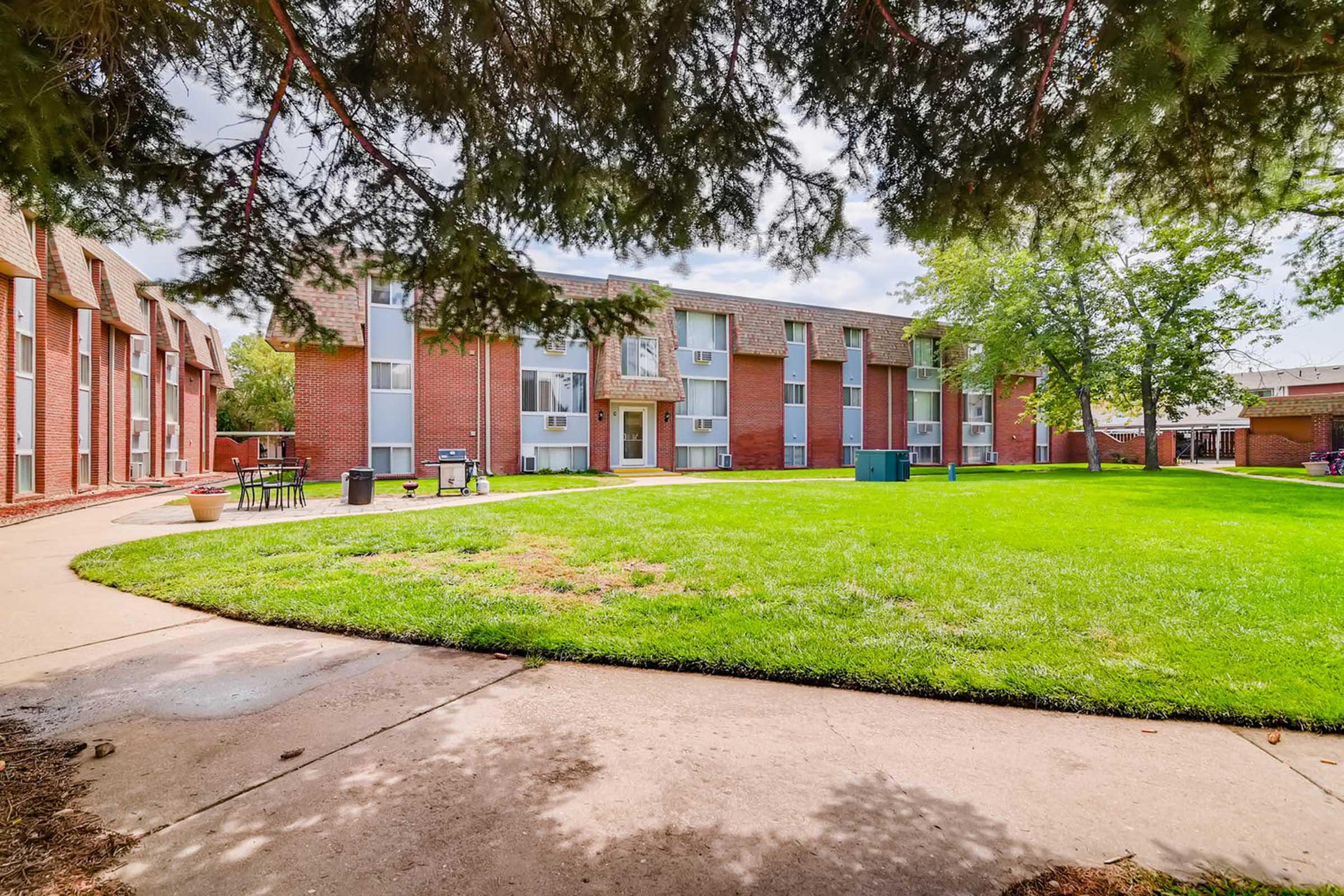A view of a brick apartment complex surrounded by a green lawn and trees. The scene includes a walkway curving around the grass, with benches and tables set up nearby. The sky is partly cloudy, adding to the pleasant, inviting atmosphere of the outdoor space.