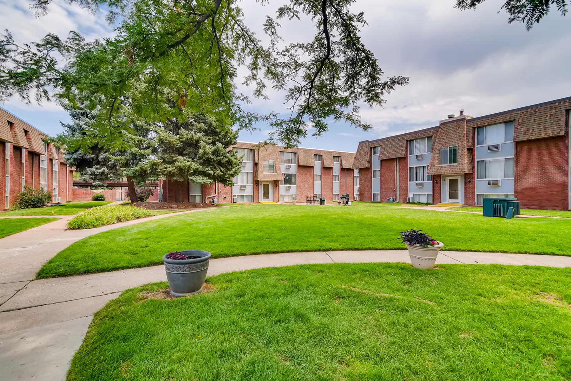 A well-maintained courtyard of an apartment complex featuring green lawns, walking paths, and landscaped areas. Several buildings with multiple units surround the space, with trees providing shade and planters adding decorative elements. The sky is overcast, suggesting a cloudy day.
