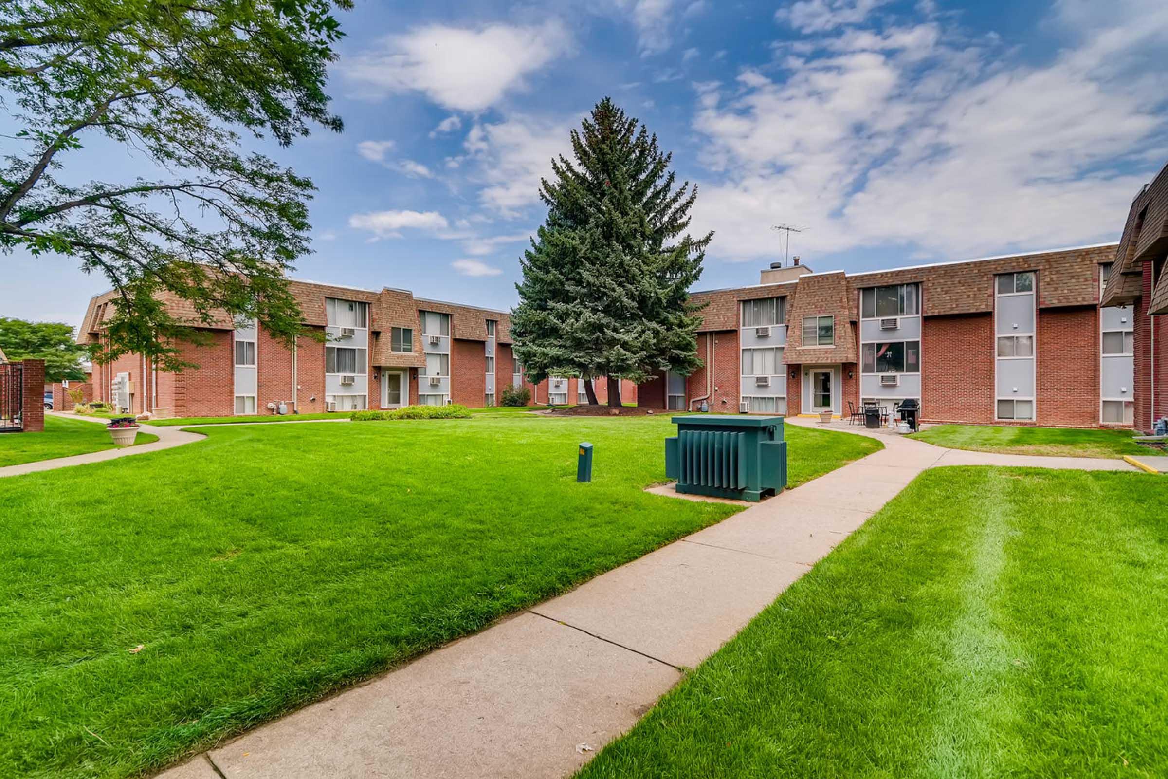 A well-maintained apartment complex surrounded by lush green grass, featuring two-story brick buildings. In the center, a tall pine tree provides shade, while pathways lead through the grassy courtyard. A green trash bin is visible along the pathway, under a partly cloudy blue sky.