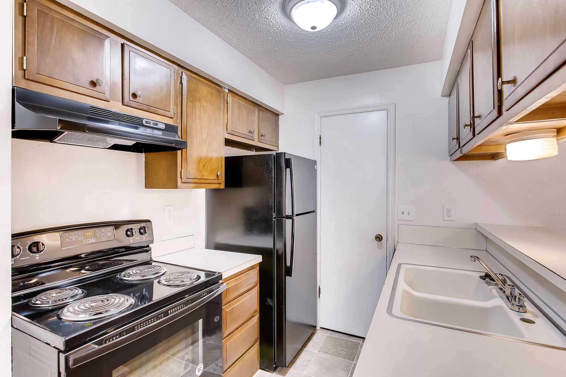 A compact kitchen featuring wooden cabinets, a black refrigerator, a stove with an oven, and a sink. The walls are light-colored, and there is a ceiling light fixture. The layout shows a functional space with essential appliances and storage, suitable for cooking and food preparation.