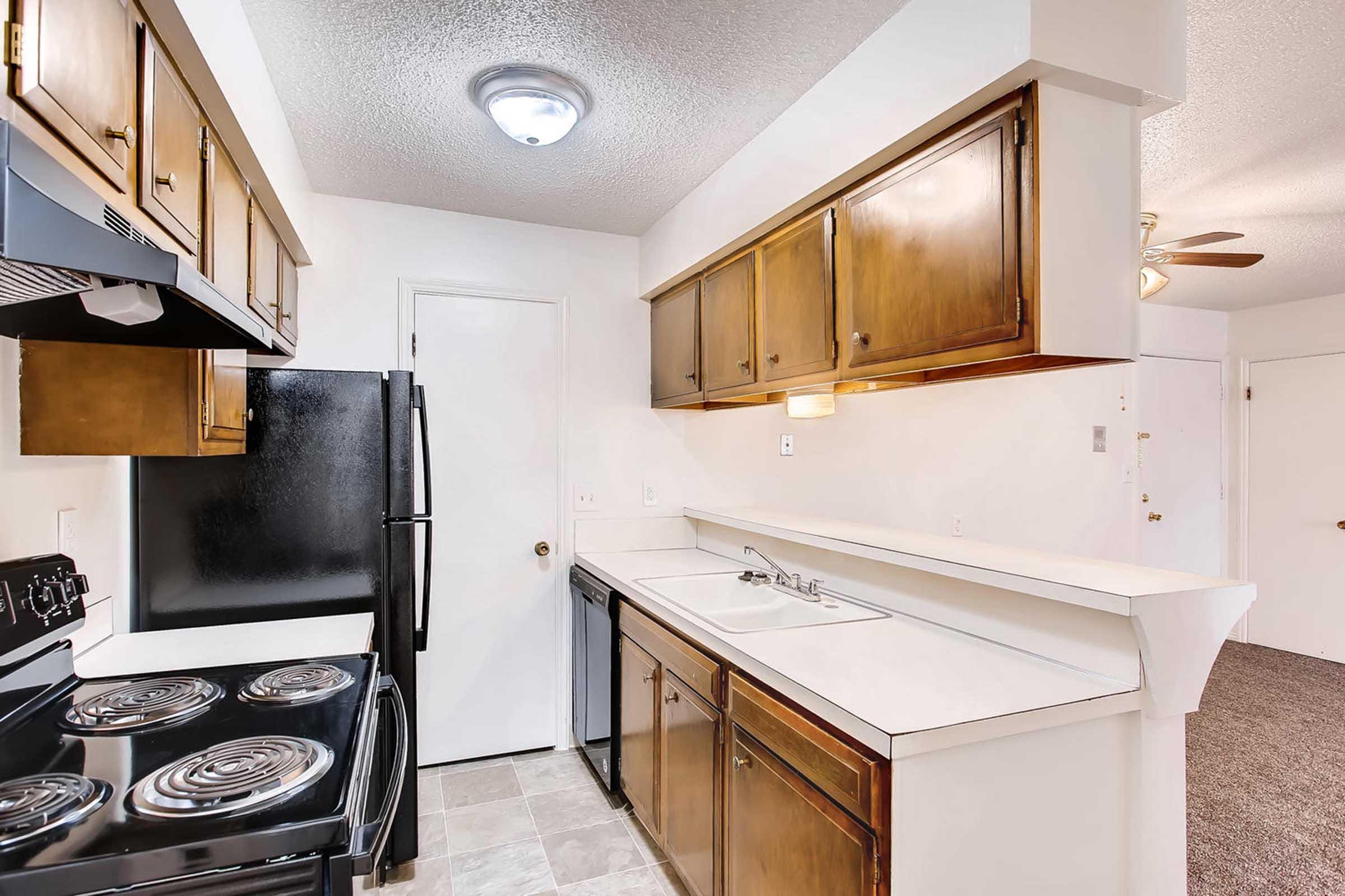 Interior of a kitchen featuring wooden cabinetry, a black refrigerator, an electric stove with an oven, and a sink with a countertop. The walls are light-colored, and a door leads to another room. The flooring appears to be tiled, with a textured ceiling and overhead lighting.
