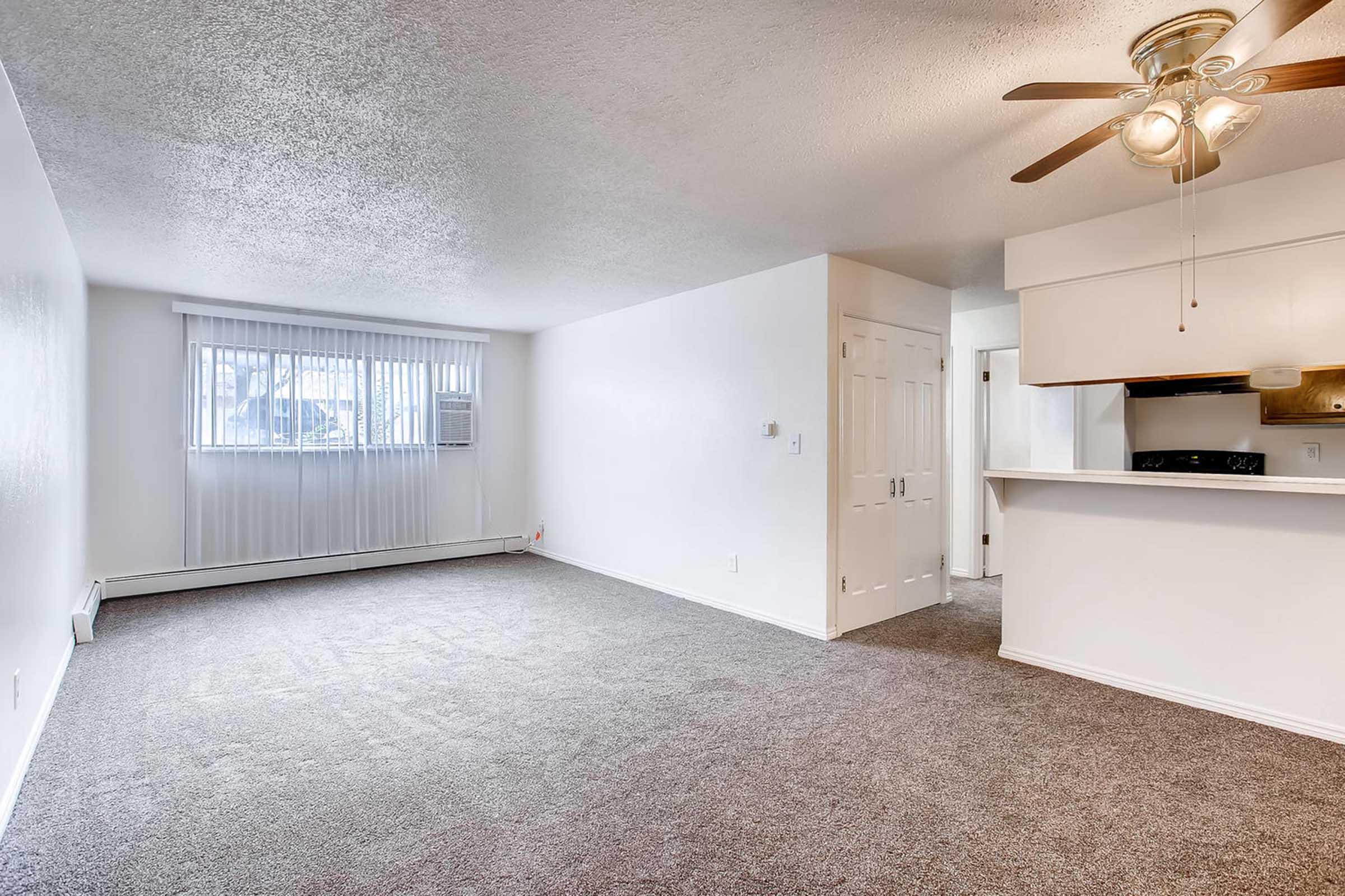 A spacious, well-lit living room featuring light gray carpet flooring, a ceiling fan, and a window with vertical blinds. The room opens to a small kitchen area with a bar counter. The walls are painted white, creating a bright and airy atmosphere.