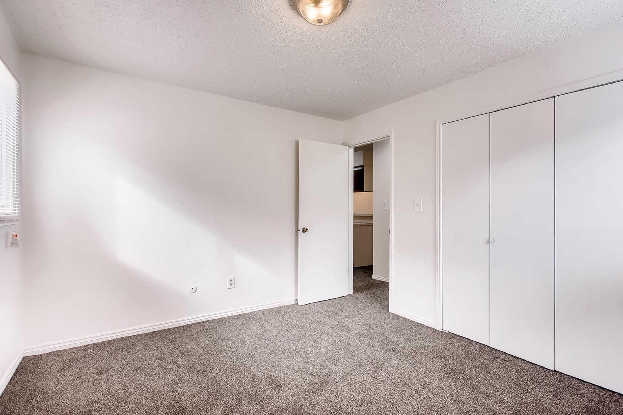 A clean, empty room with beige carpet and white walls. A door is slightly ajar on the right, leading to another space. Natural light comes through a window, casting soft shadows. The room has a ceiling light fixture, providing a bright and inviting atmosphere.