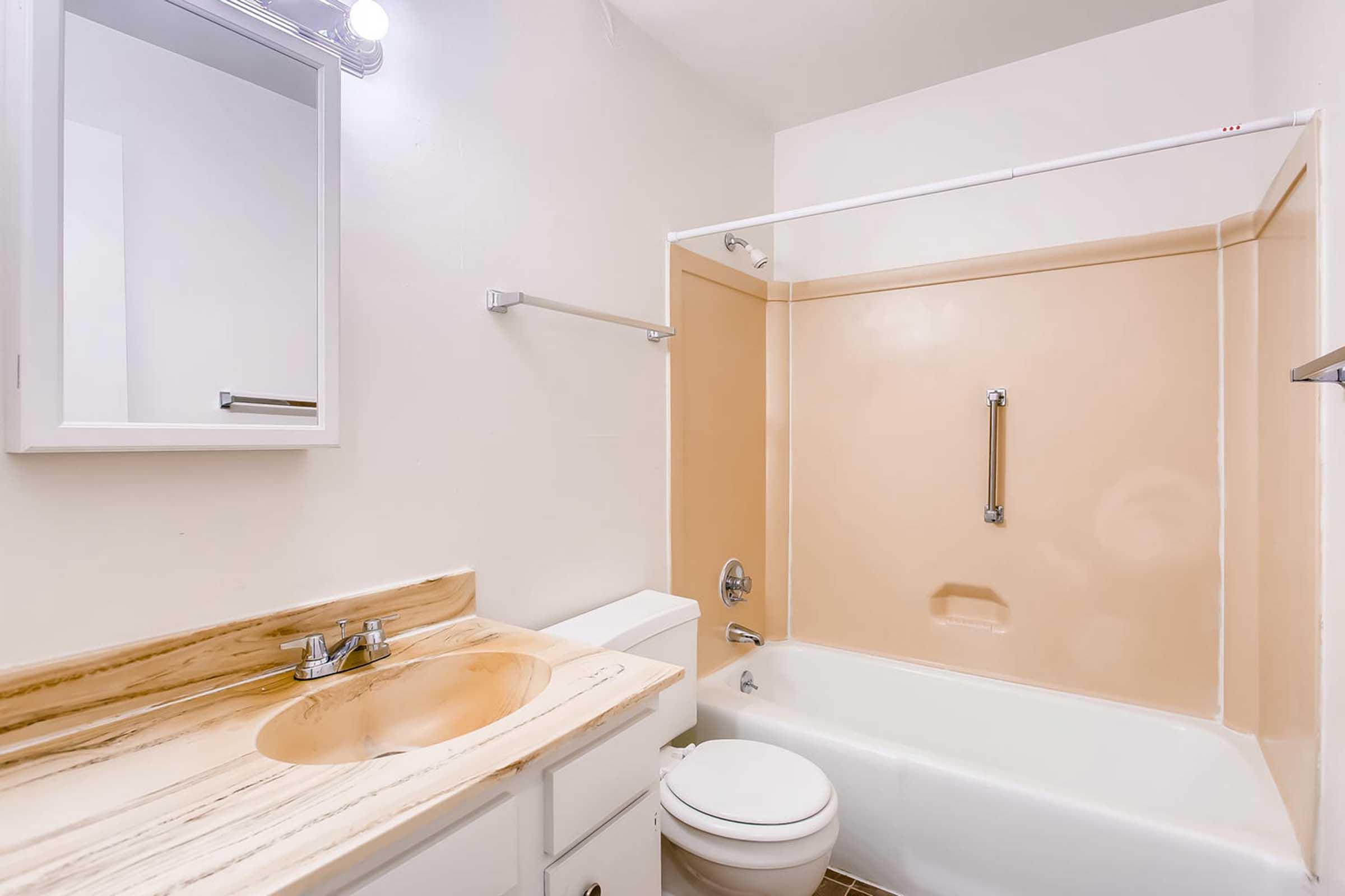 A clean, well-lit bathroom featuring a beige bathtub with a shower, a white toilet, and a small sink with a marble countertop. The walls are painted light colors, and there's a mirror above the sink. Chrome fixtures are present, giving a modern touch to the space.