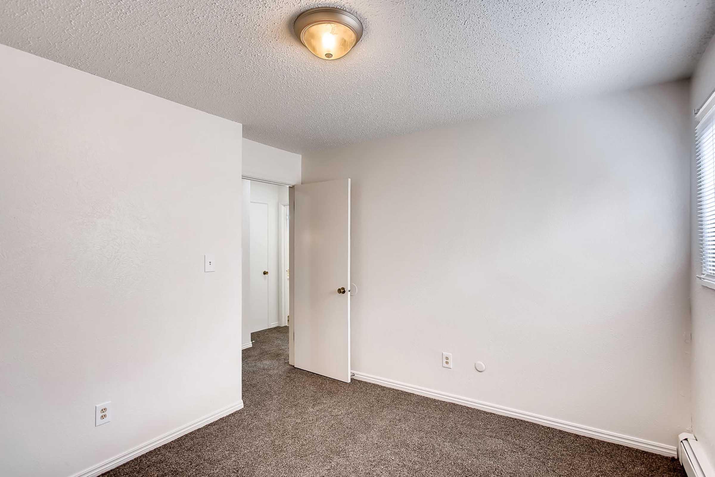 A small, minimalistic room with beige walls and carpet. A light fixture on the ceiling provides soft illumination. An open door on the left leads to another space, while a window on the right allows natural light. The room is empty, featuring no furniture or decorations, creating a clean and simple atmosphere.