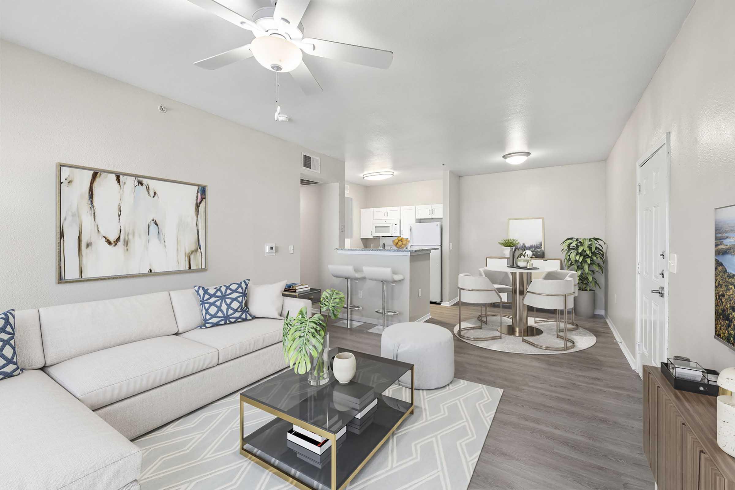 A modern living room featuring a light-colored sectional sofa, a coffee table with plant decor, and a circular area rug. The space includes a dining area with a small round table and chairs, and a kitchen visible in the background with white cabinetry. Soft lighting and neutral tones create a welcoming atmosphere.