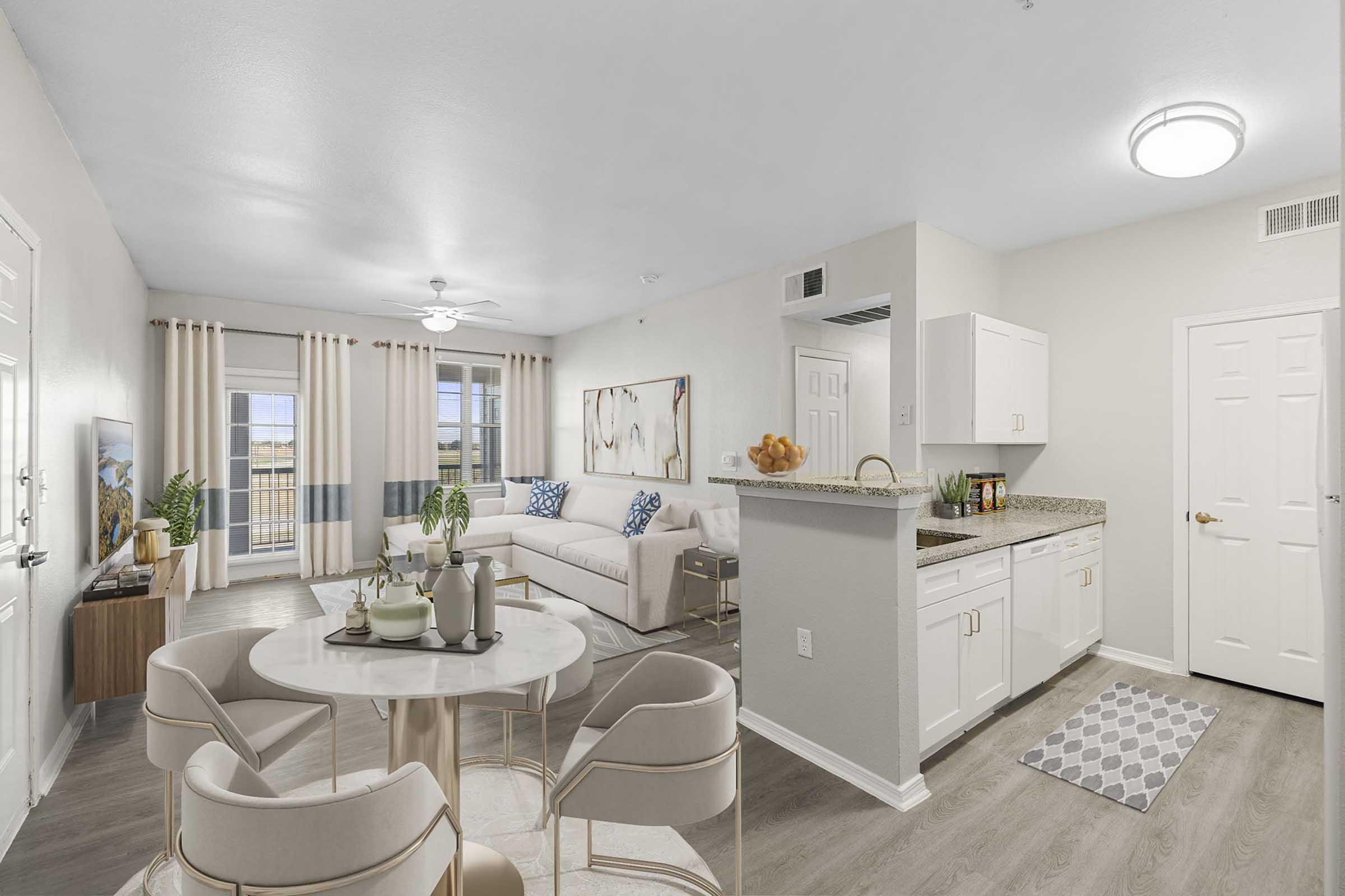 Modern apartment interior featuring a bright living room with a large sofa, decorative wall art, and a TV. A dining area with a round table and stylish chairs is visible, alongside a kitchen with white cabinets and granite countertops. Natural light fills the space from large windows.