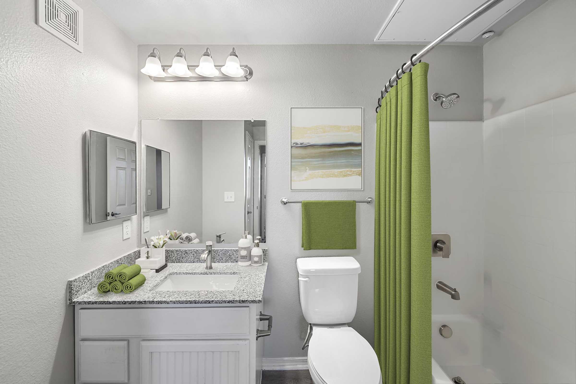 A clean bathroom featuring a white wall, a granite countertop sink with rolled green towels, a mirror, and a shower with a green curtain. A small framed abstract painting and a green hand towel are also visible, contributing to a fresh and modern aesthetic.