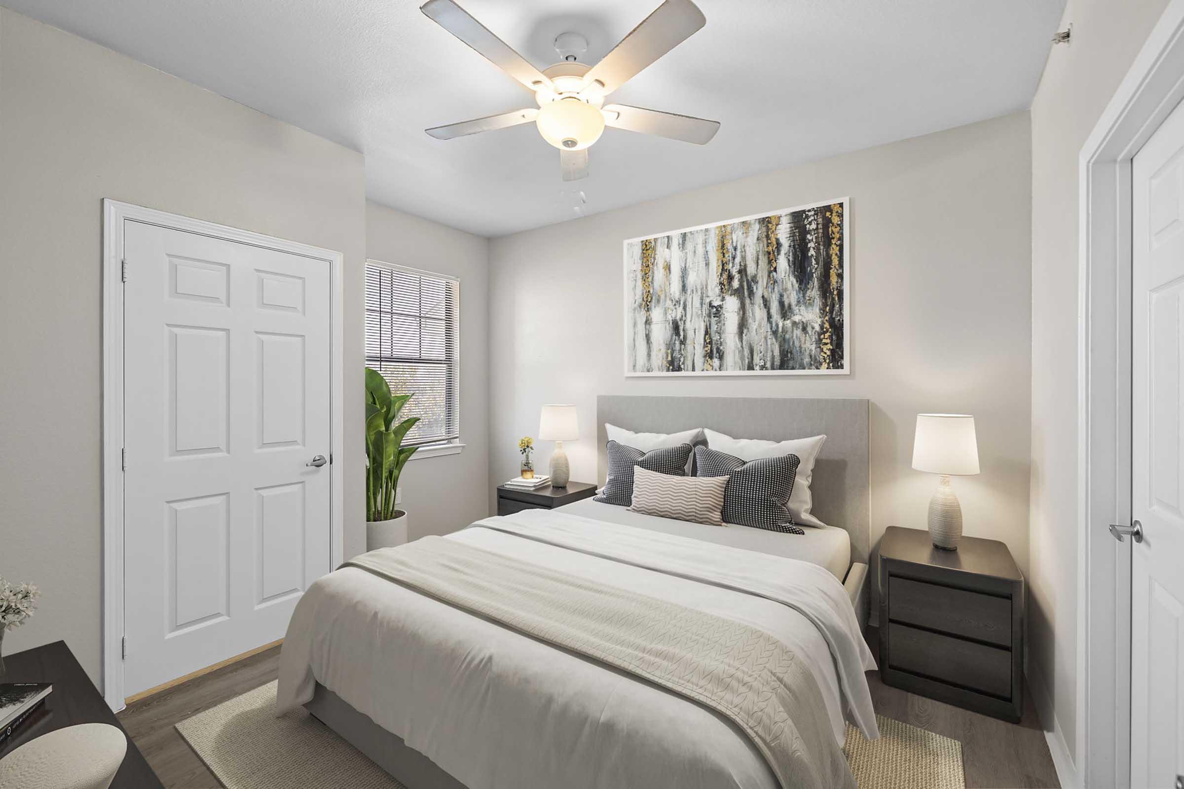 A cozy bedroom featuring a queen-sized bed with gray and white bedding. There's a decorative painting above the bed, two bedside tables with lamps, and a potted plant near the window. The walls are painted in neutral tones, and the room has a ceiling fan for added comfort.