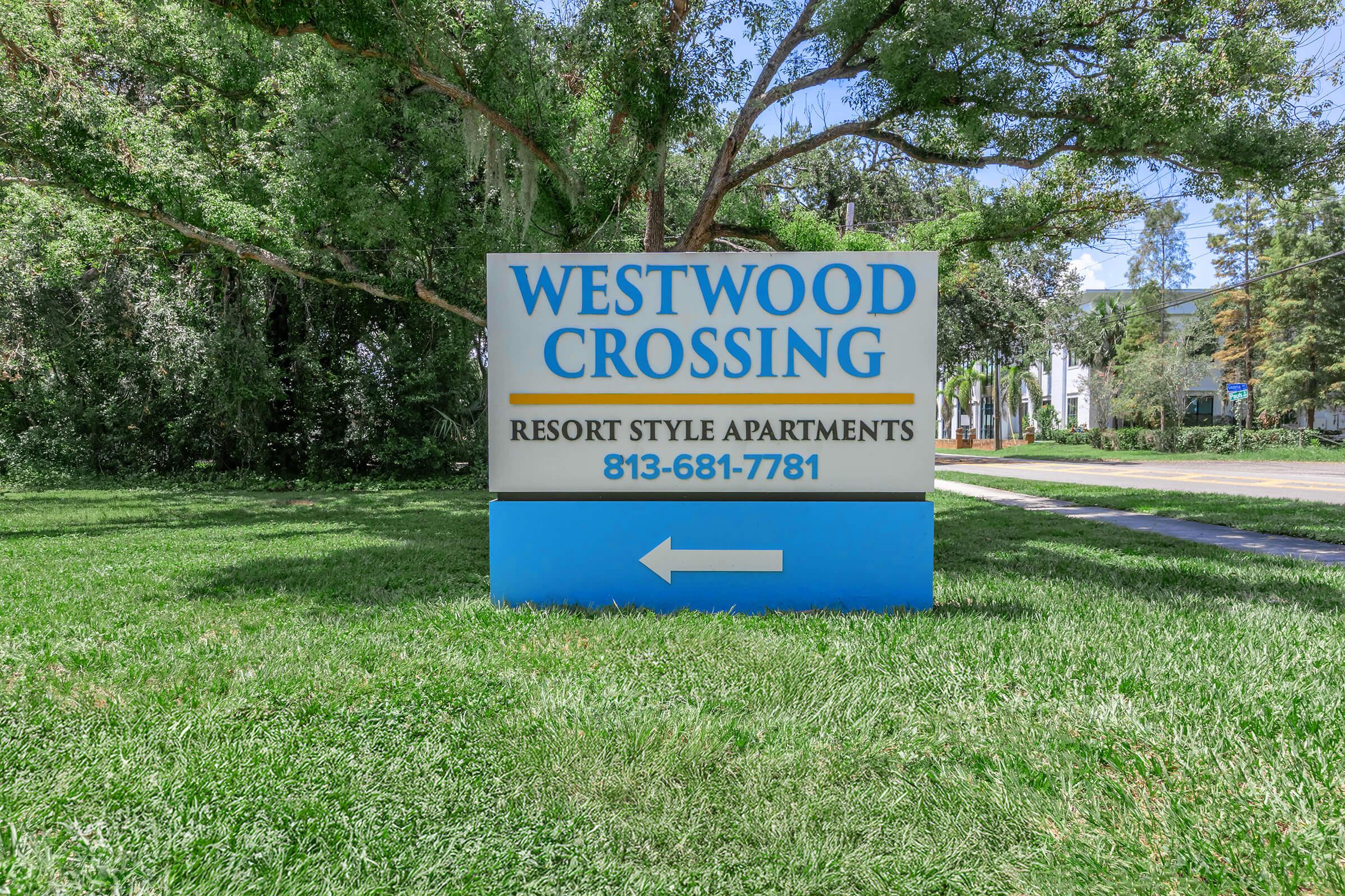 Sign for Westwood Crossing Resort Style Apartments featuring the name "WESTWOOD CROSSING" at the top with a phone number, 813-681-7781, below. An arrow points to the left, with a green grassy area and trees in the background.