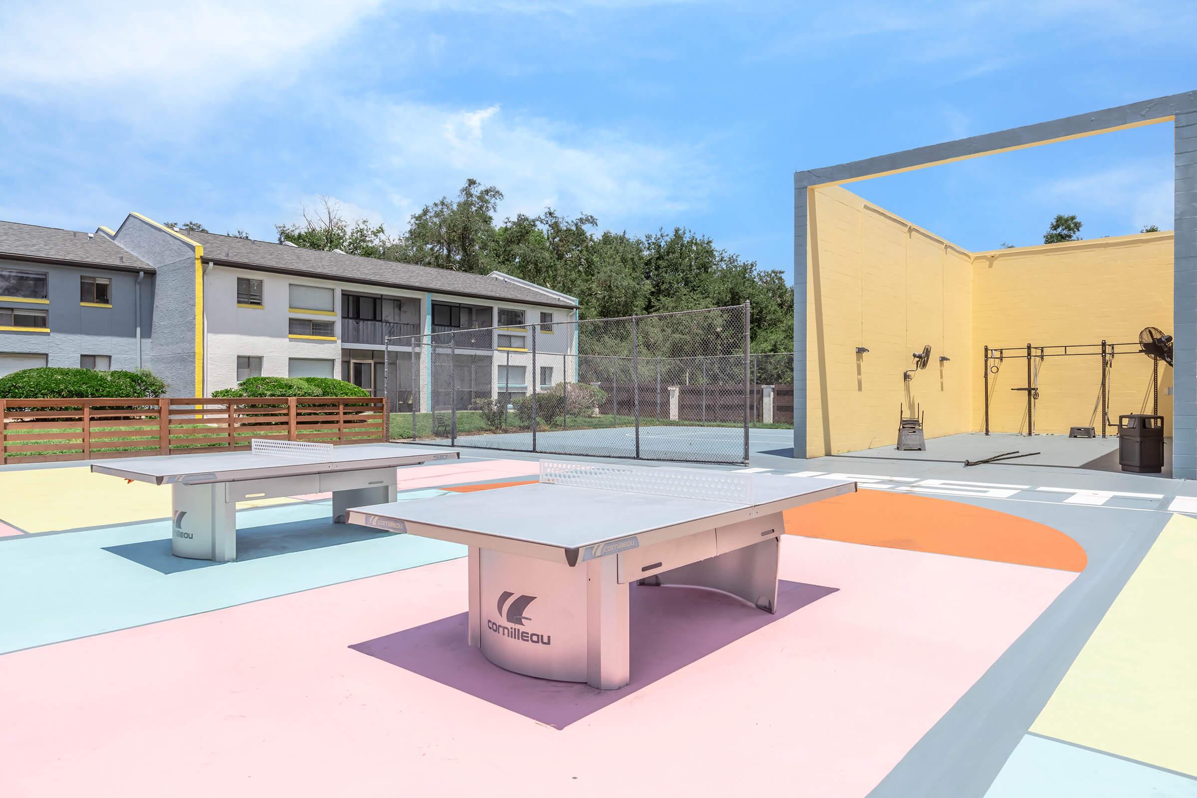 A vibrant outdoor recreational area featuring two metallic ping pong tables on a colorful pavement, surrounded by grassy areas and residential buildings in the background. A light blue sky with a few clouds is visible above, enhancing the cheerful atmosphere of the space.