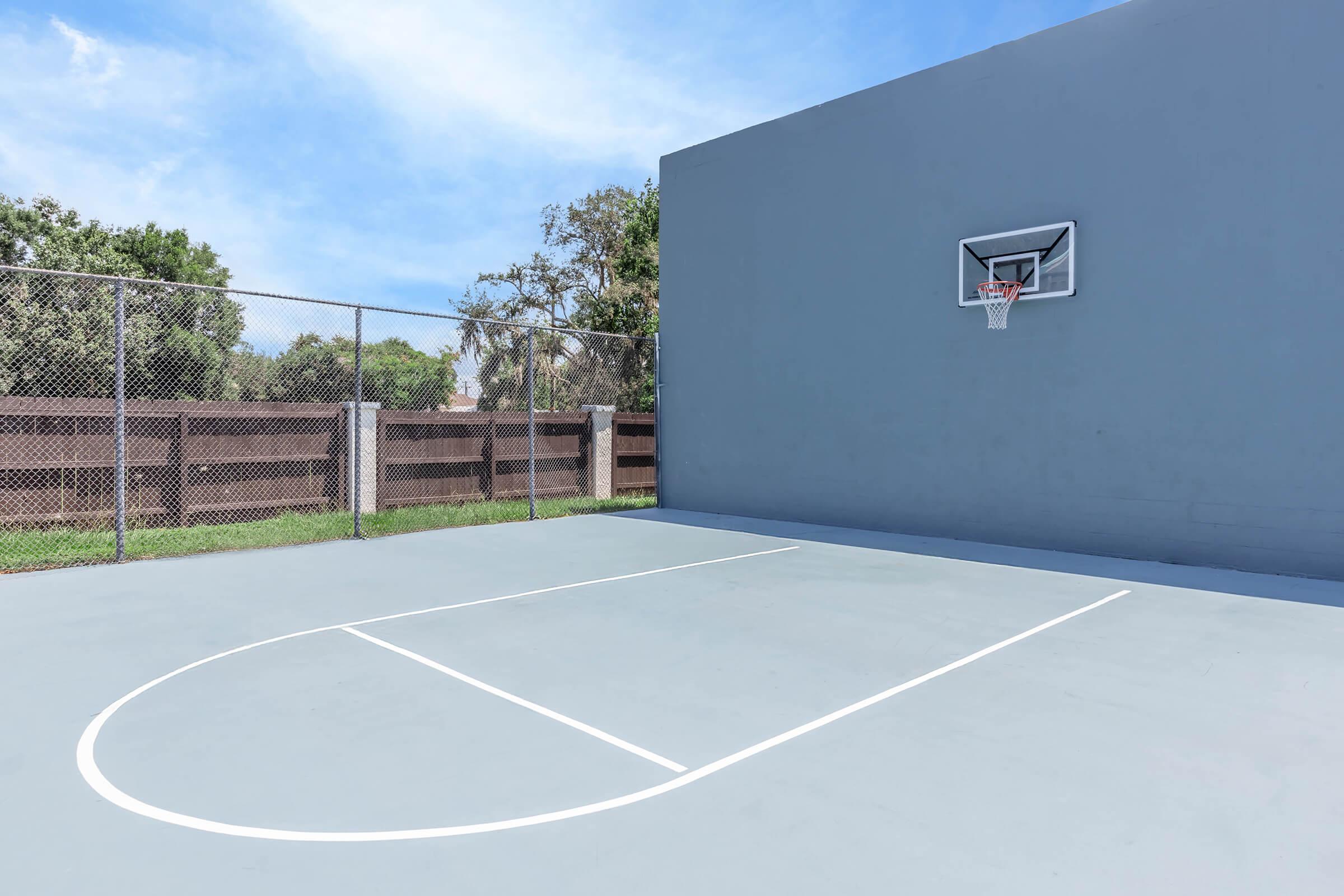 A basketball court featuring a gray surface with a single hoop mounted on a gray wall. The court is enclosed by a chain-link fence, surrounded by greenery and a wooden fence in the background, under a clear blue sky.
