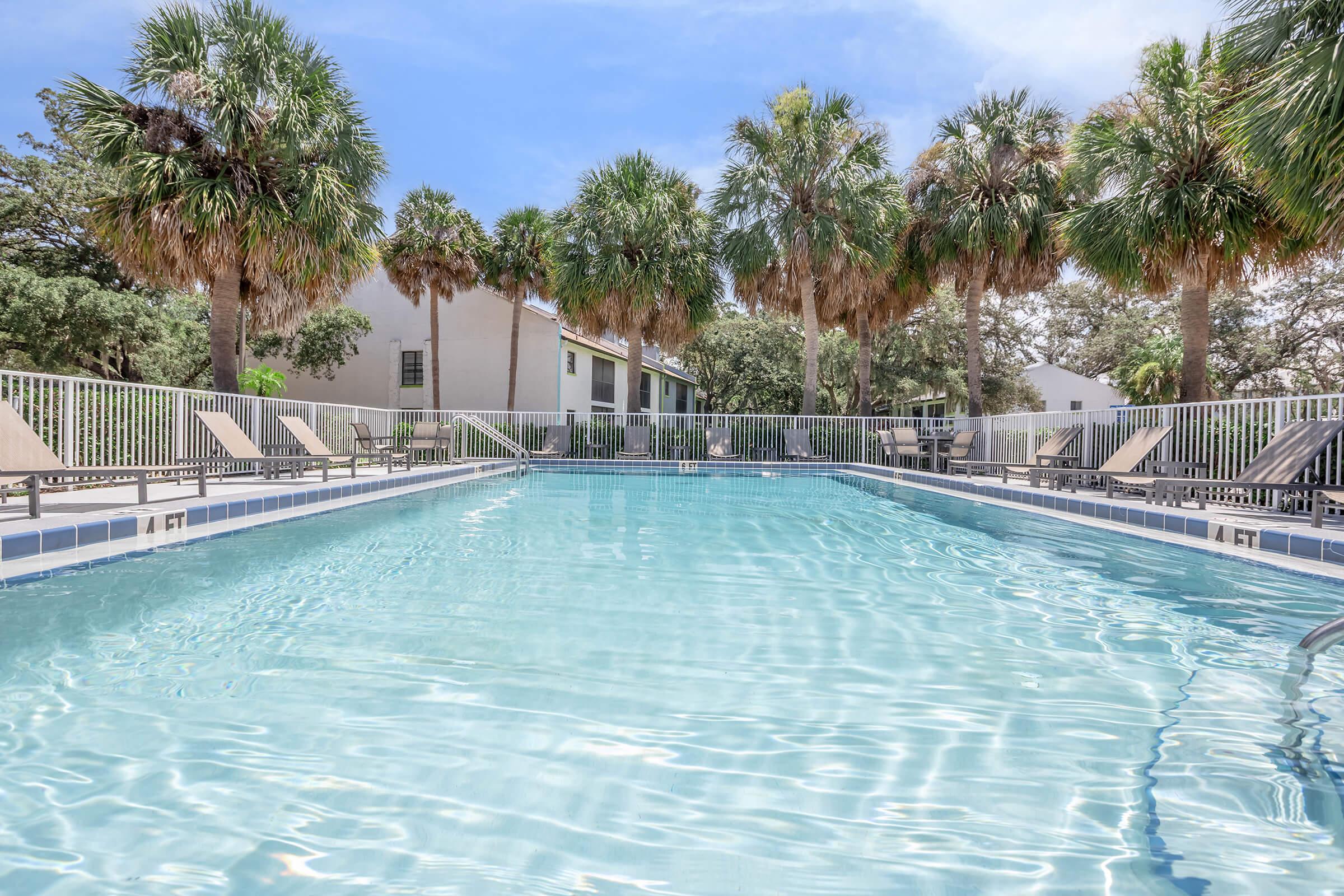 A clear swimming pool surrounded by palm trees and lounge chairs. The pool's surface reflects the sky, and a residential building is visible in the background, partially shaded by the trees. The scene conveys a relaxed, tropical atmosphere.