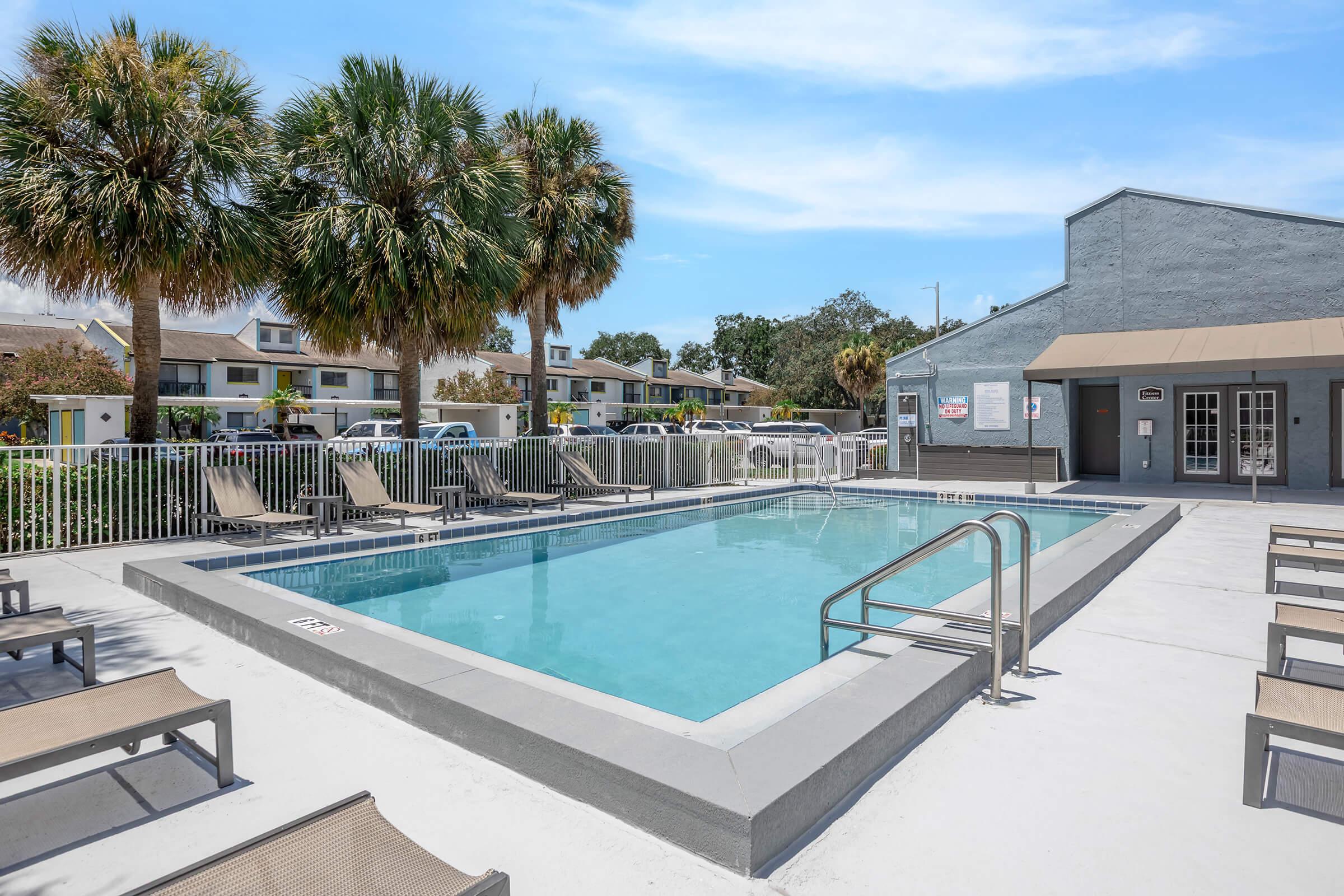 A clear swimming pool surrounded by lounge chairs, palm trees, and a building in the background. The pool area is sunlit with a blue sky overhead, creating a relaxing outdoor leisure space.