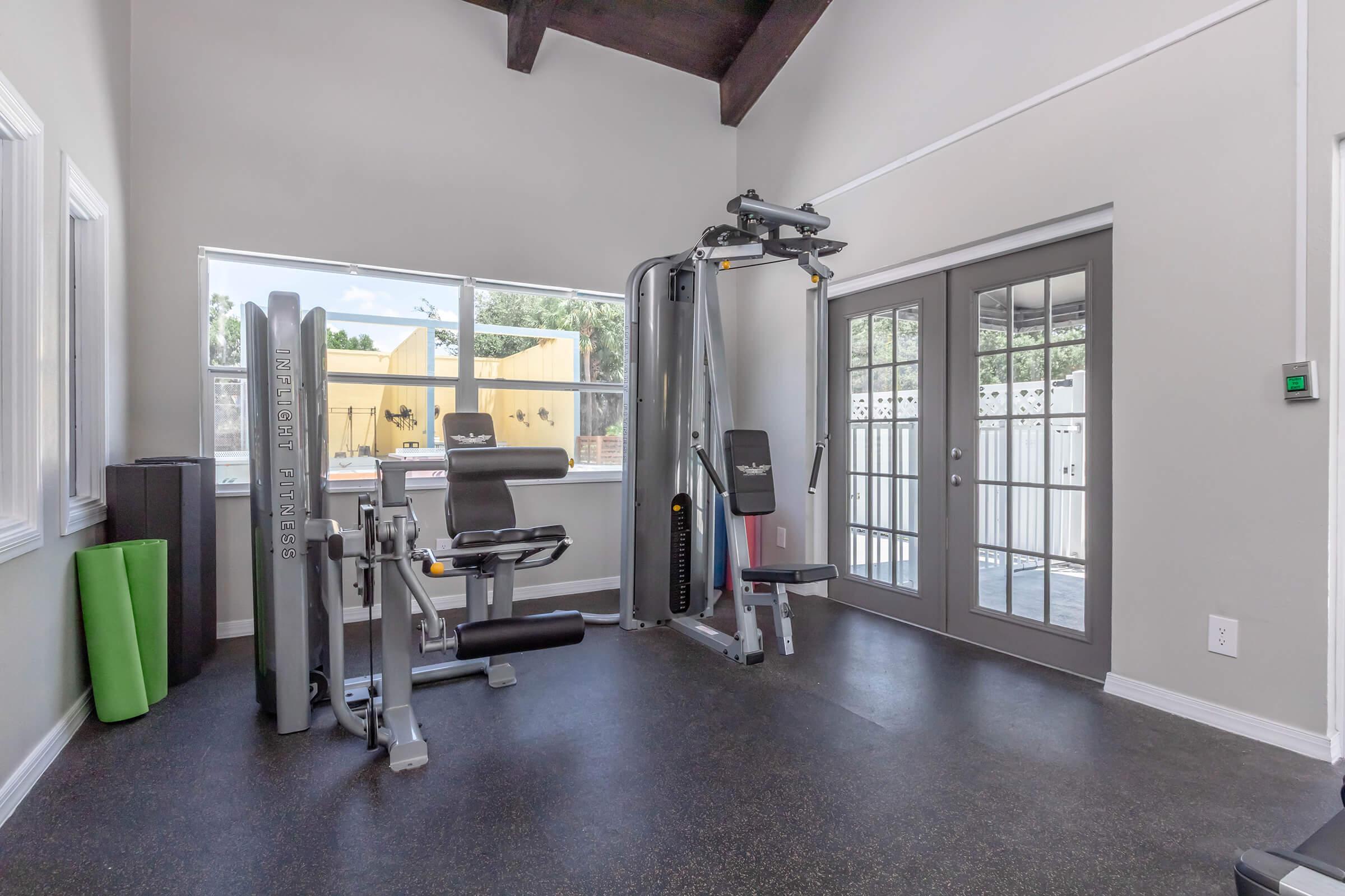 A well-lit home gym featuring exercise equipment including a multi-gym machine, a weight bench, and green foam rollers. Large windows allow natural light to fill the space, and a door leads to an outdoor area. The flooring is dark and the walls are painted a light color.